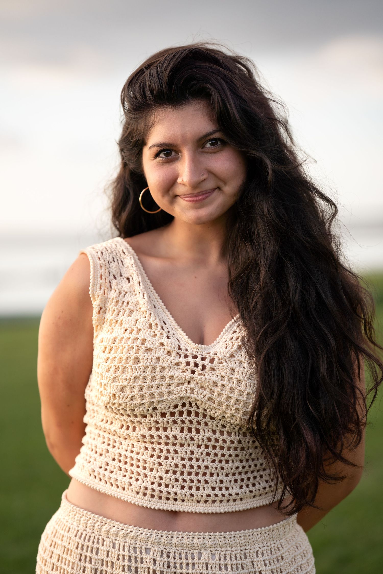 Young woman with long dark hair wearing a cream crochet crop top and skirt set, smiling outdoors near the ocean.