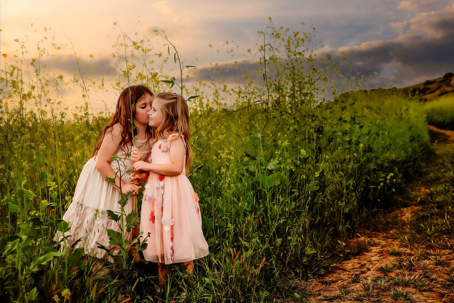 Two figures in flowing dresses share a moment together in a wildflower field at sunset.