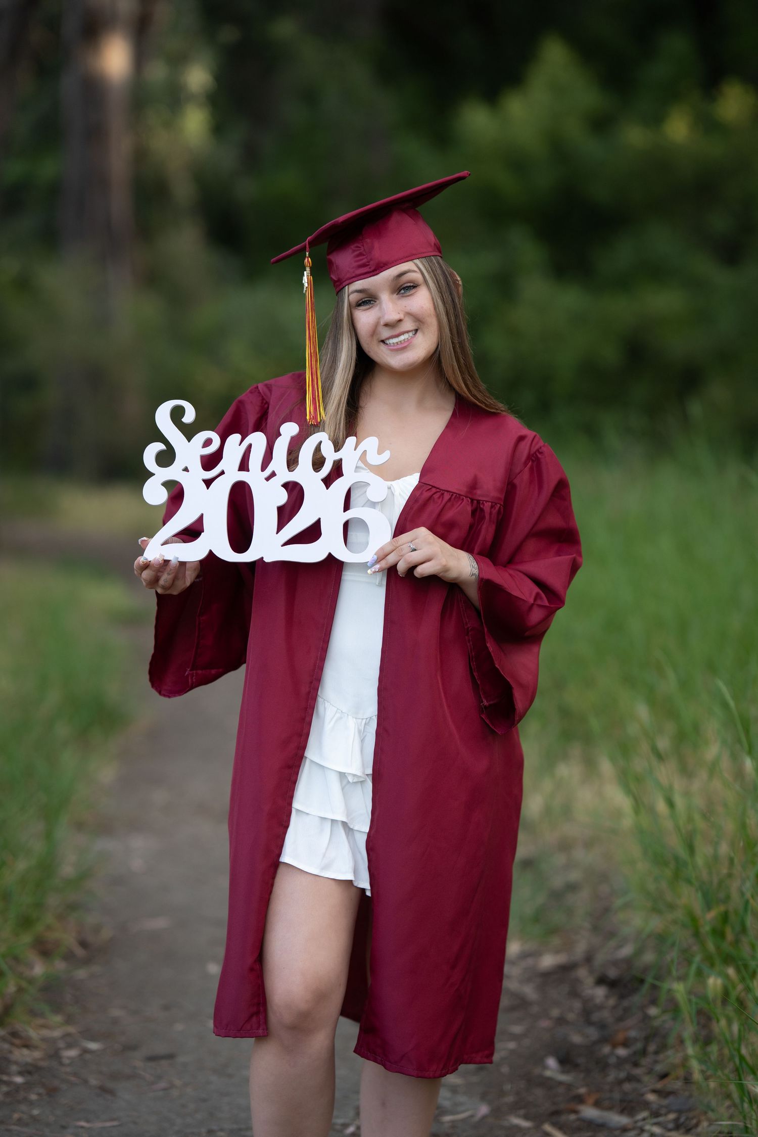 Smiling high school senior in maroon cap and gown holding a white Senior 2026 sign outdoors.