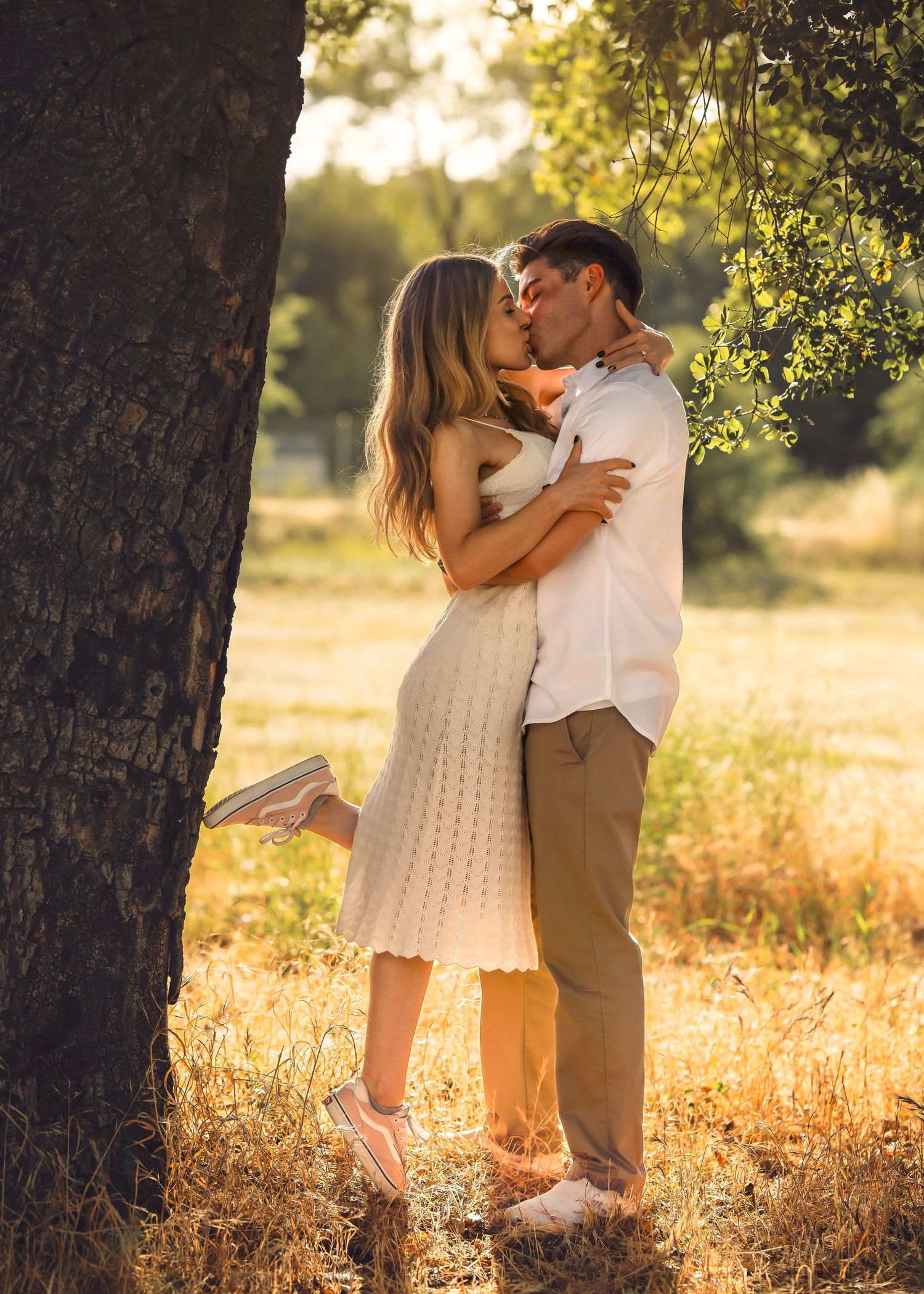 Couple kissing romantically near a tree in a sunlit field, woman in white dress, man in casual attire.