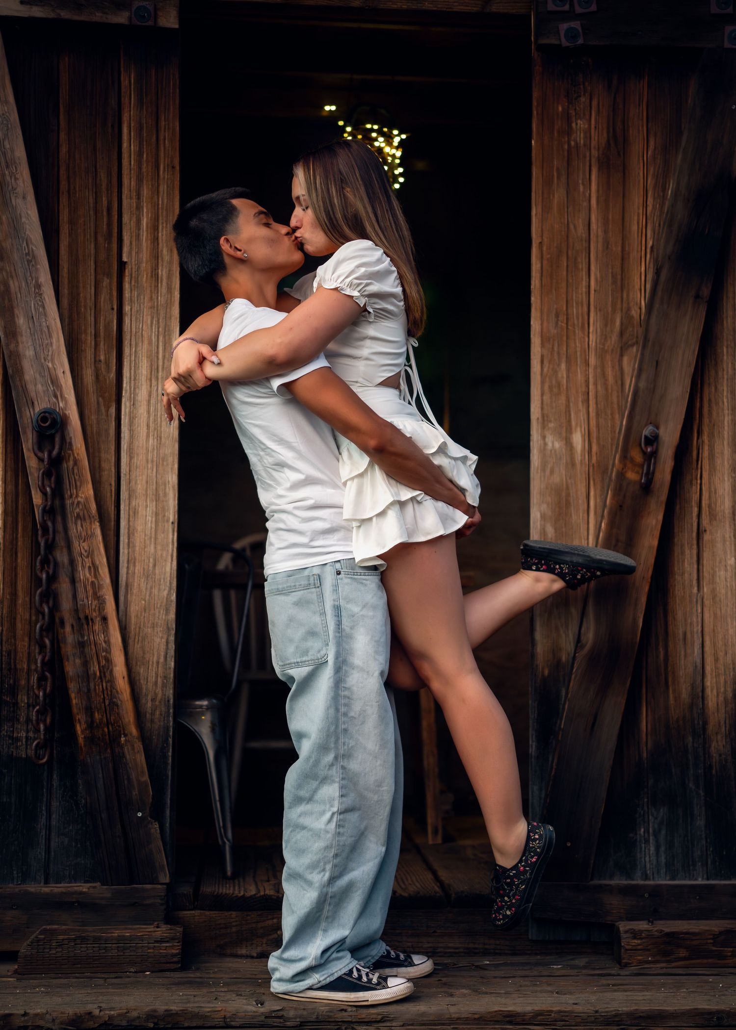 Couple sharing a romantic kiss in a rustic barn doorway, woman lifted in embrace, both wearing white tops.