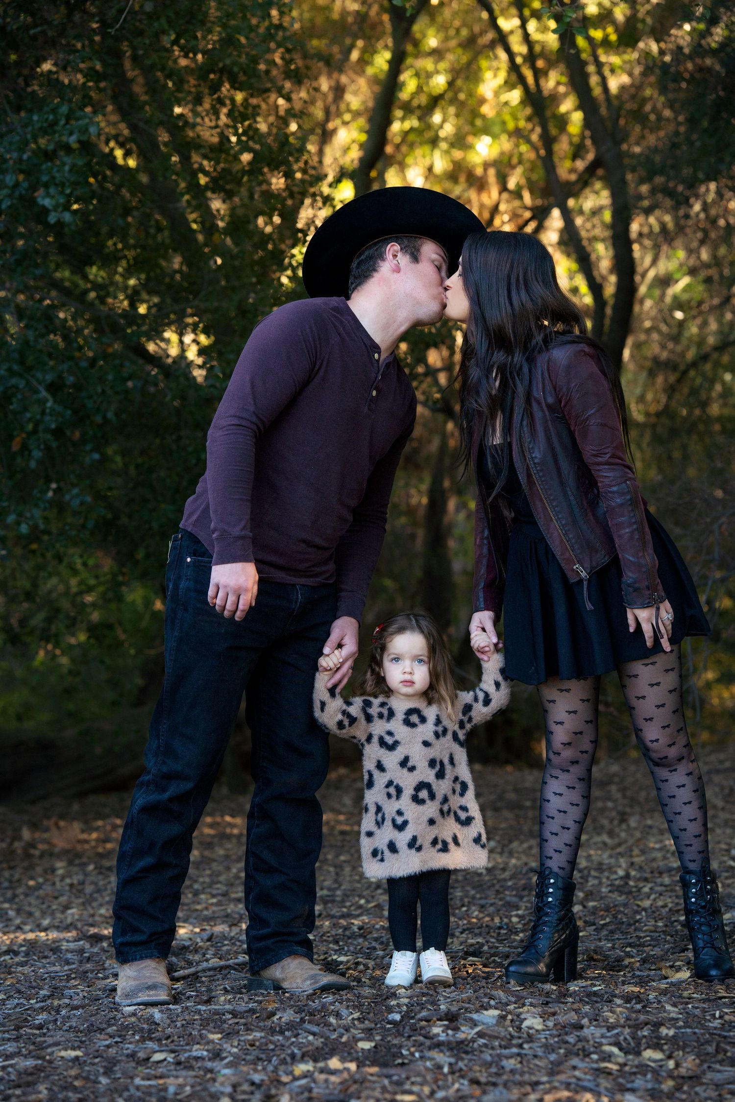 Romantic couple shares a kiss while their child stands between them on a forest path.