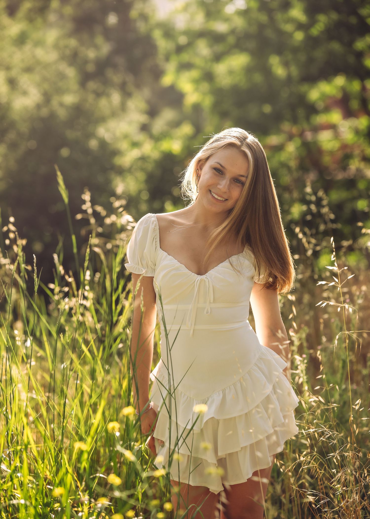 Young woman in white ruffled mini dress smiling in sunlit meadow with tall grass and trees.