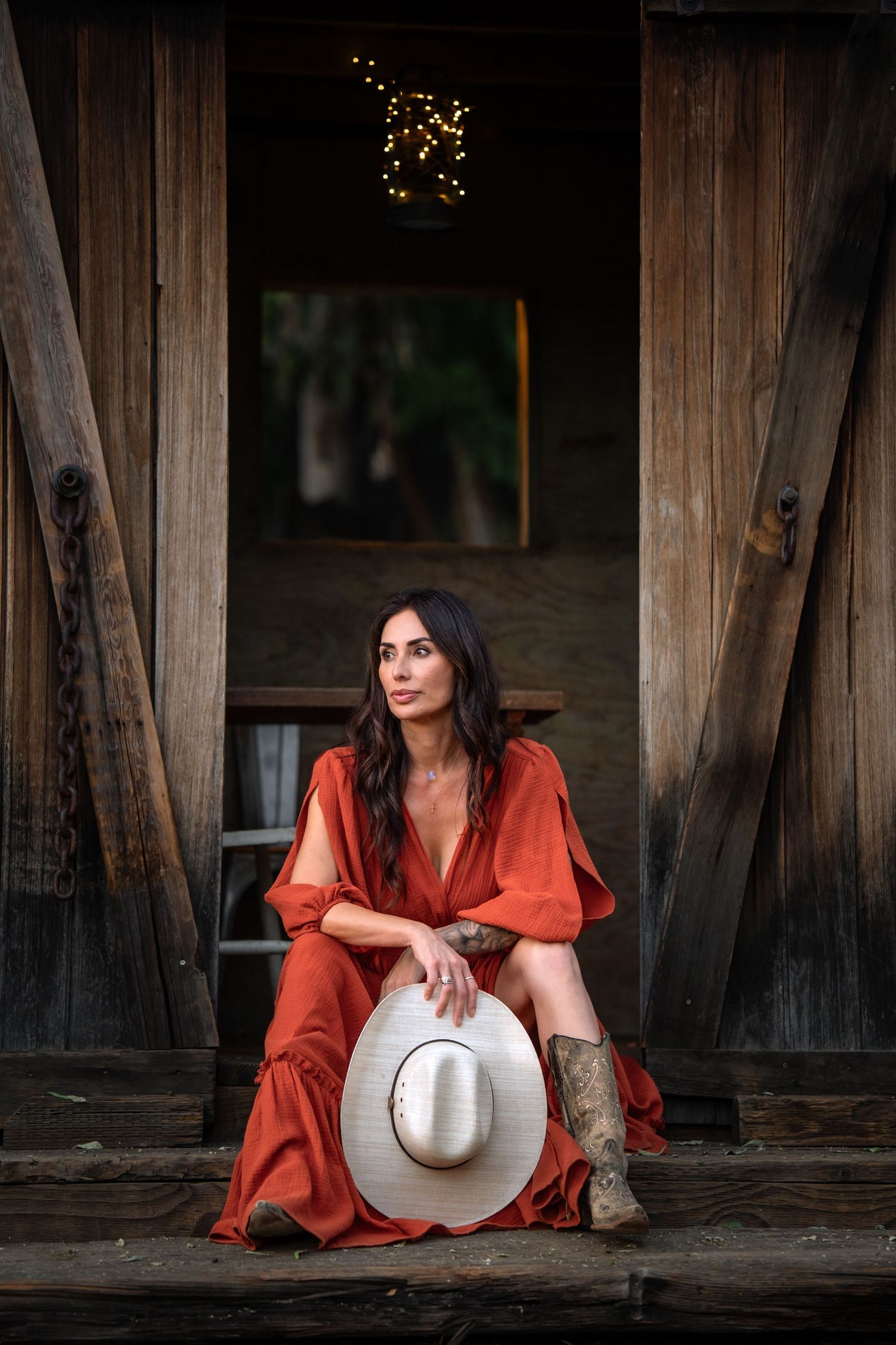 Woman in red dress and cowboy boots holding white hat sits in rustic barn doorway.