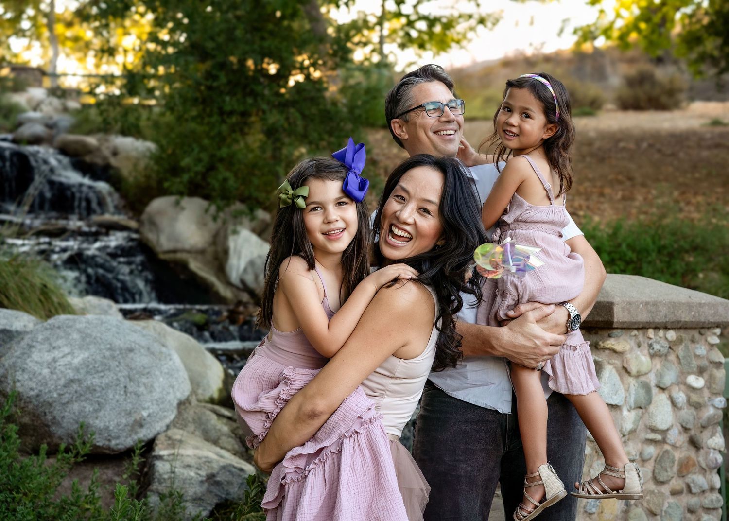 A loving family portrait taken outdoors by a waterfall with matching pink dresses and a blue butterfly accessory.
