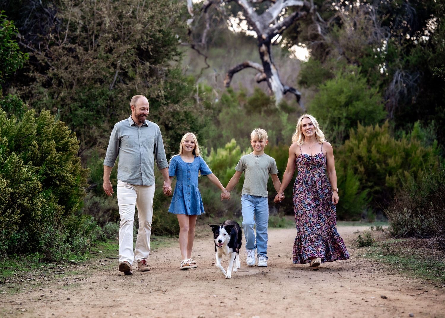 A family walks together on a dirt path with their border collie dog in a natural outdoor setting.
