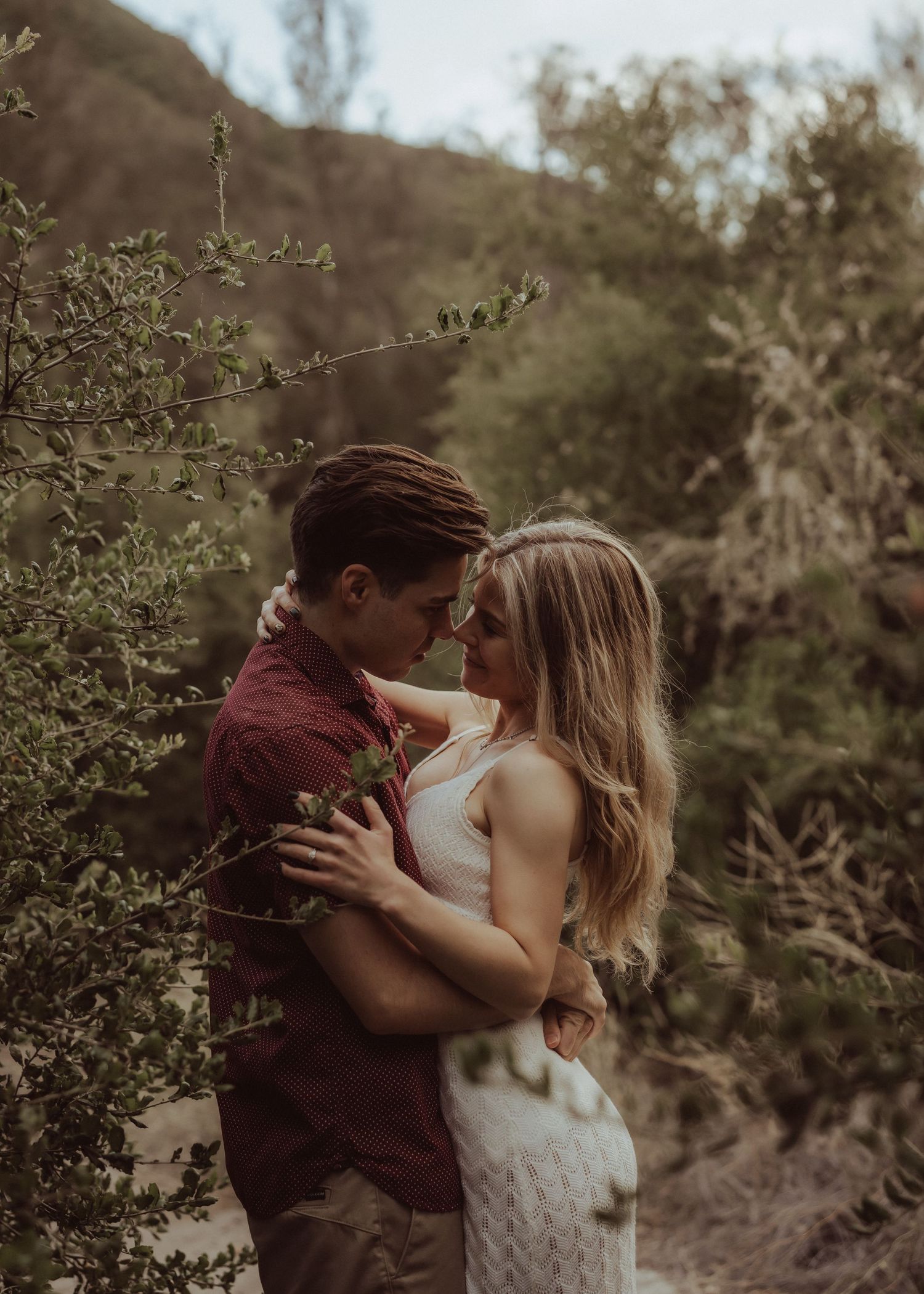 Couple embracing intimately in a lush outdoor setting with trees and mountains, wearing casual boho attire.