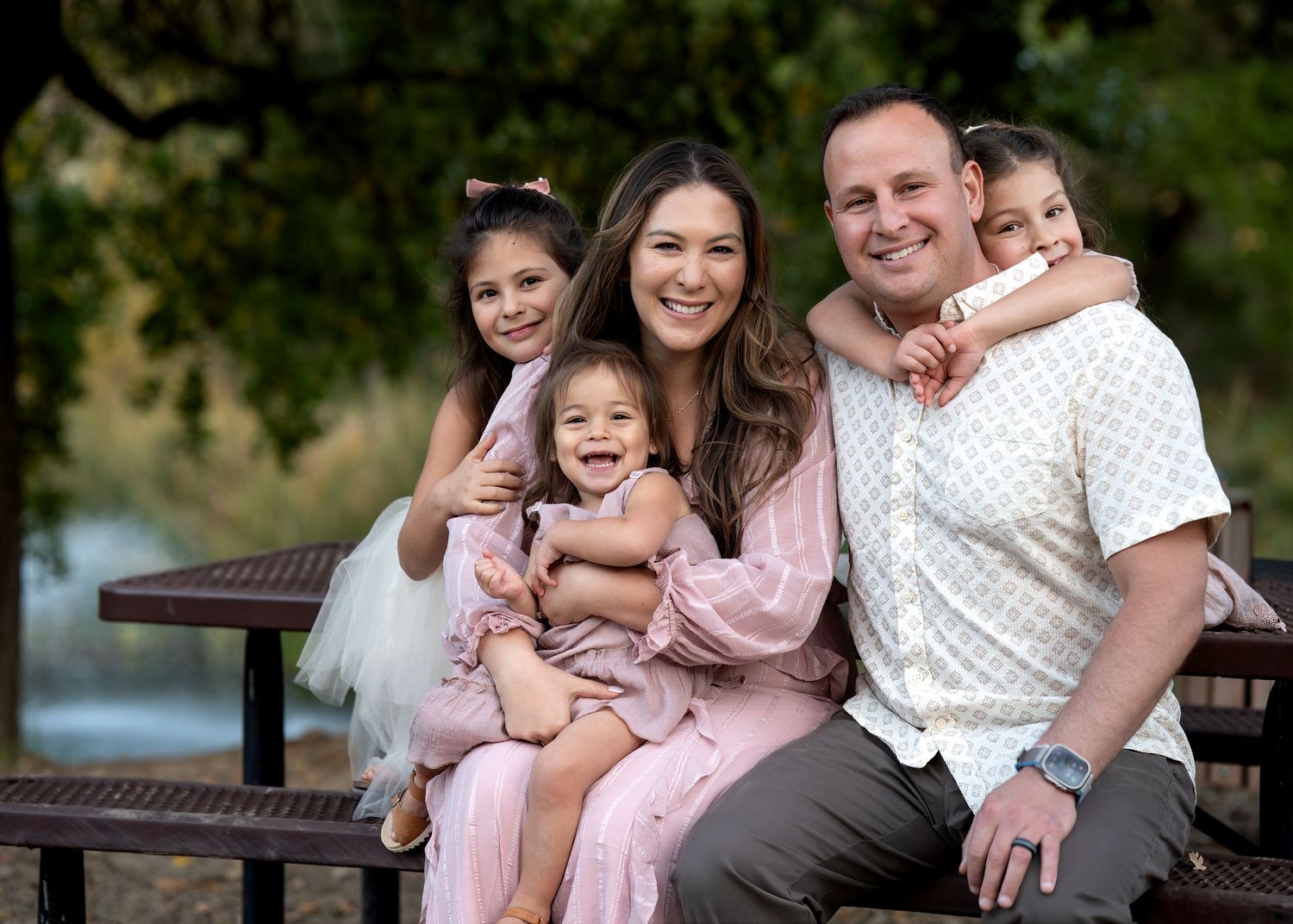 Happy family sitting together on a wooden bench near a lake in coordinated pink and white outfits.