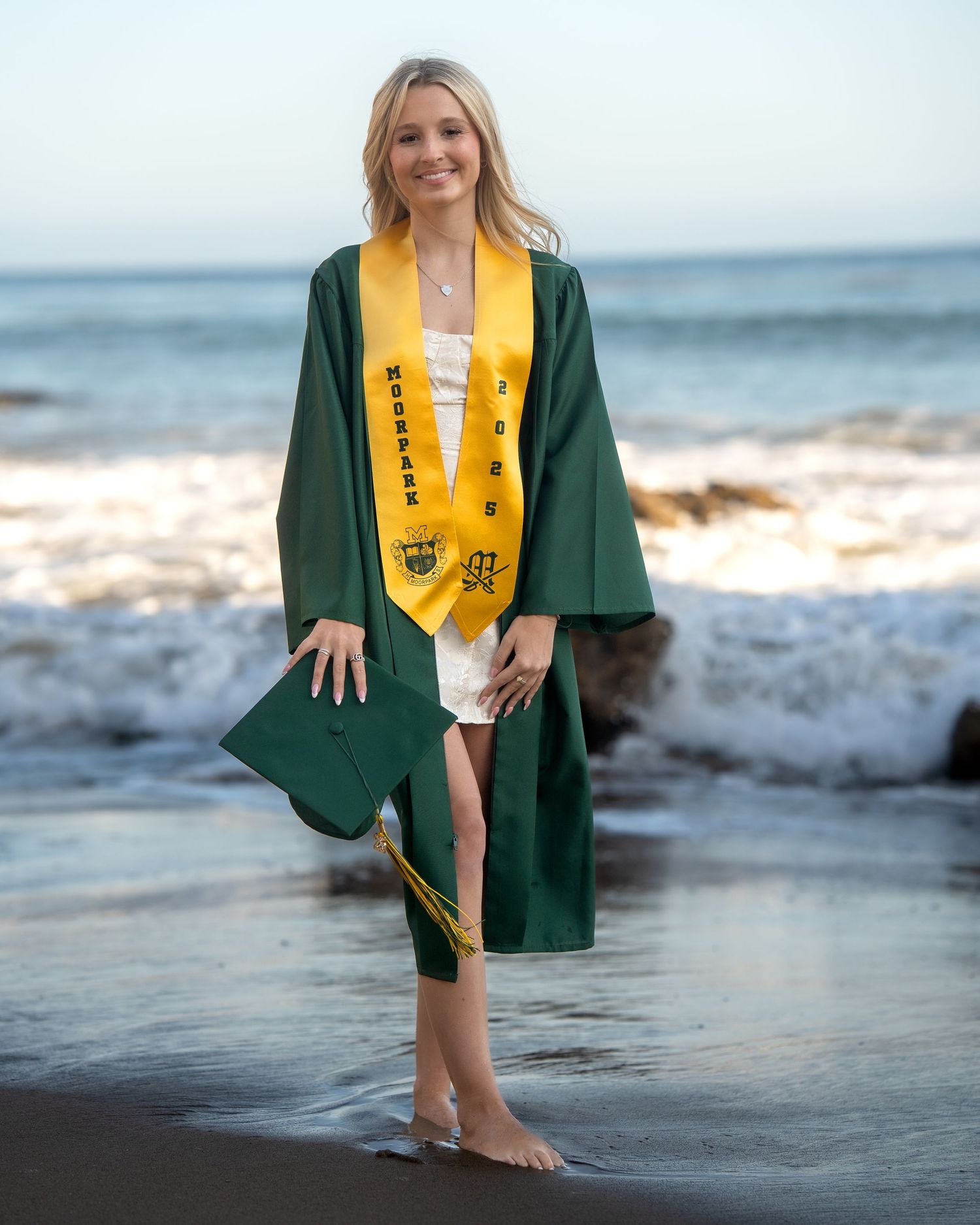 Graduate in green cap and gown poses on beach with waves in background.