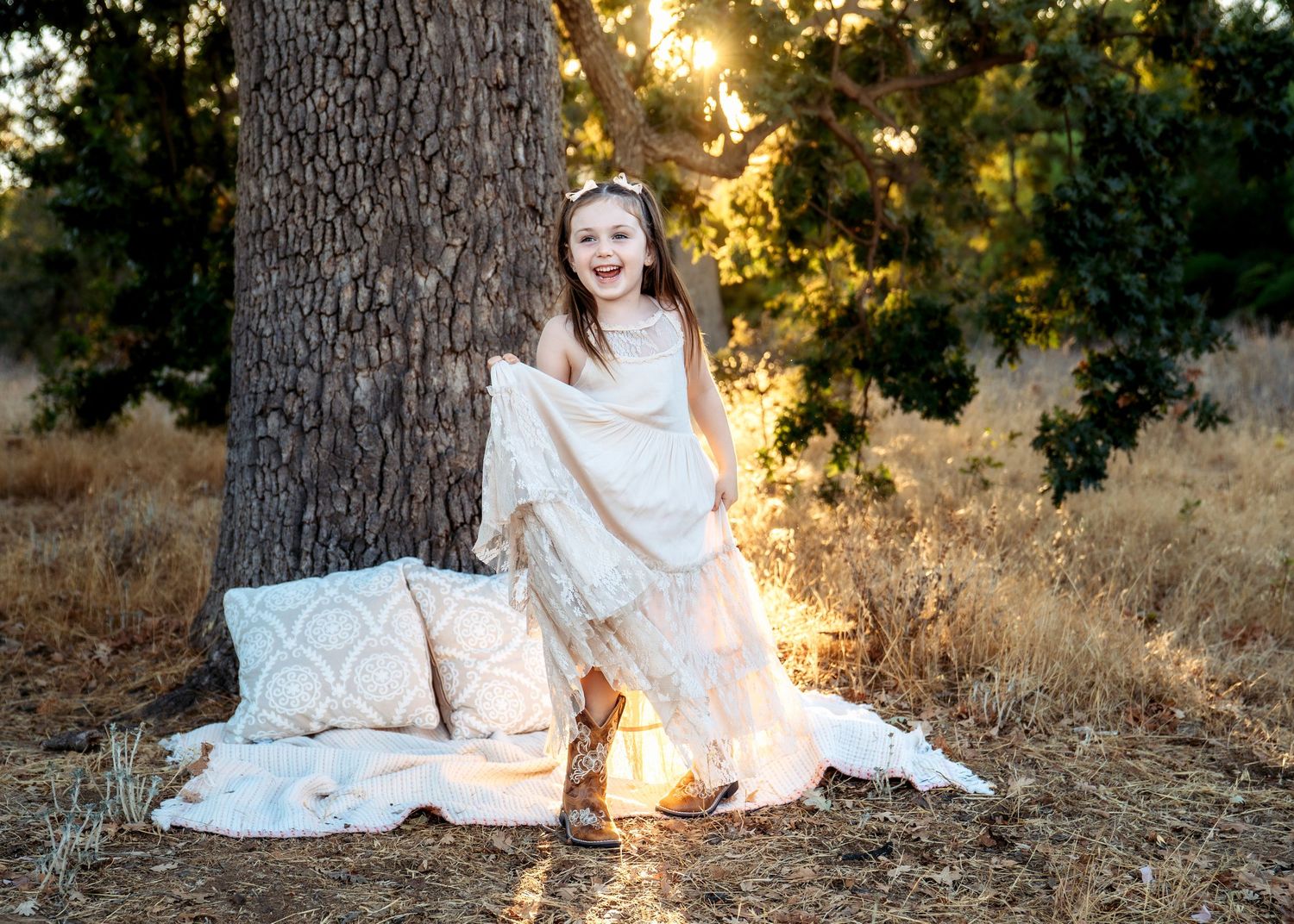 Dreamy outdoor portrait with white dress and pillows against tree trunk at sunset.