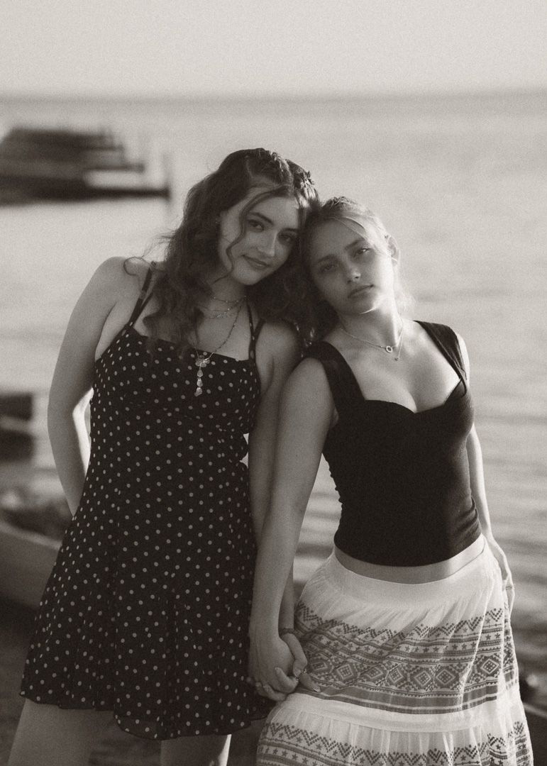 Black and white photo of two young women posing together near a waterfront, one in a polka dot dress.