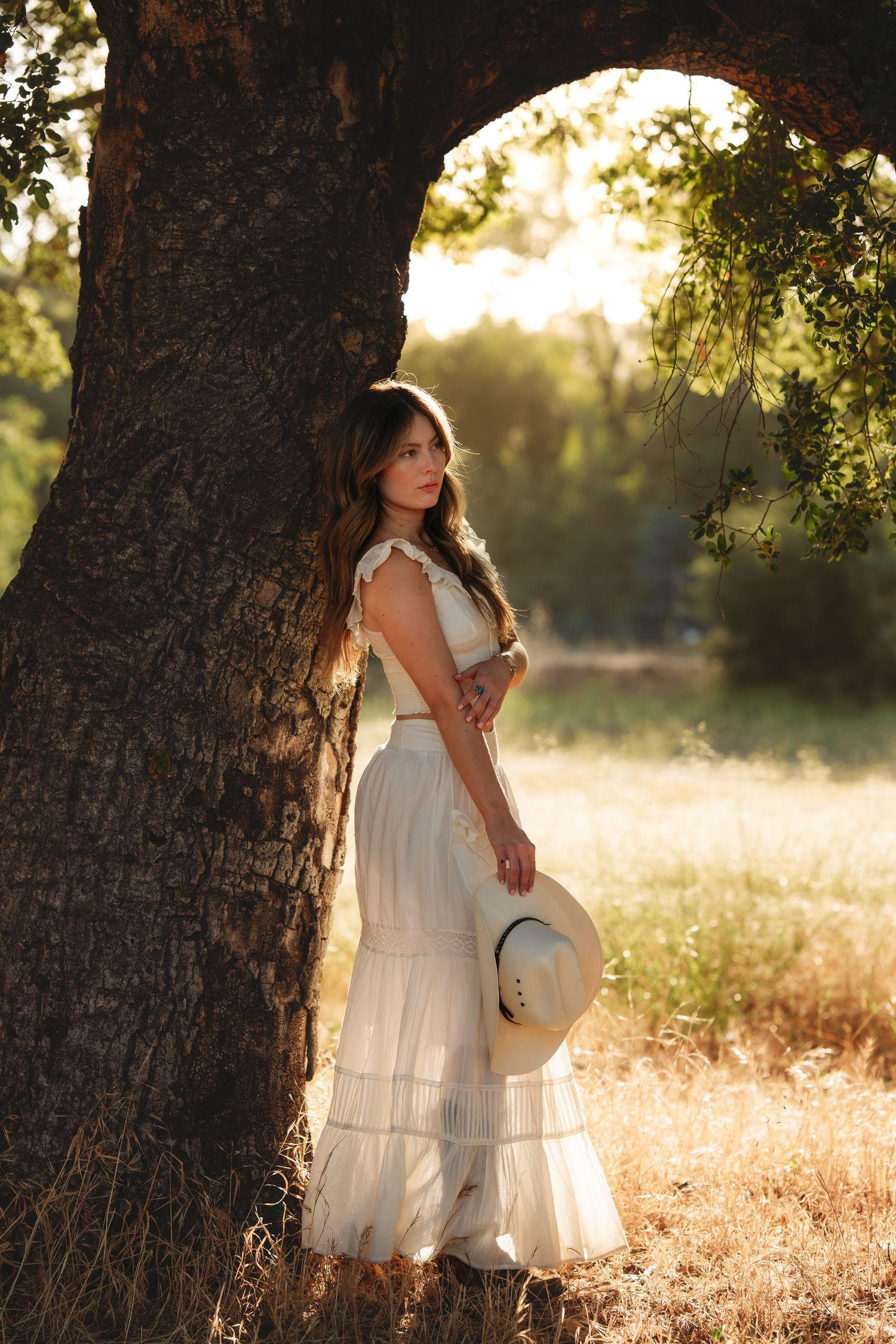 Young woman in white boho dress holding straw hat, leaning against large tree in golden sunset light.