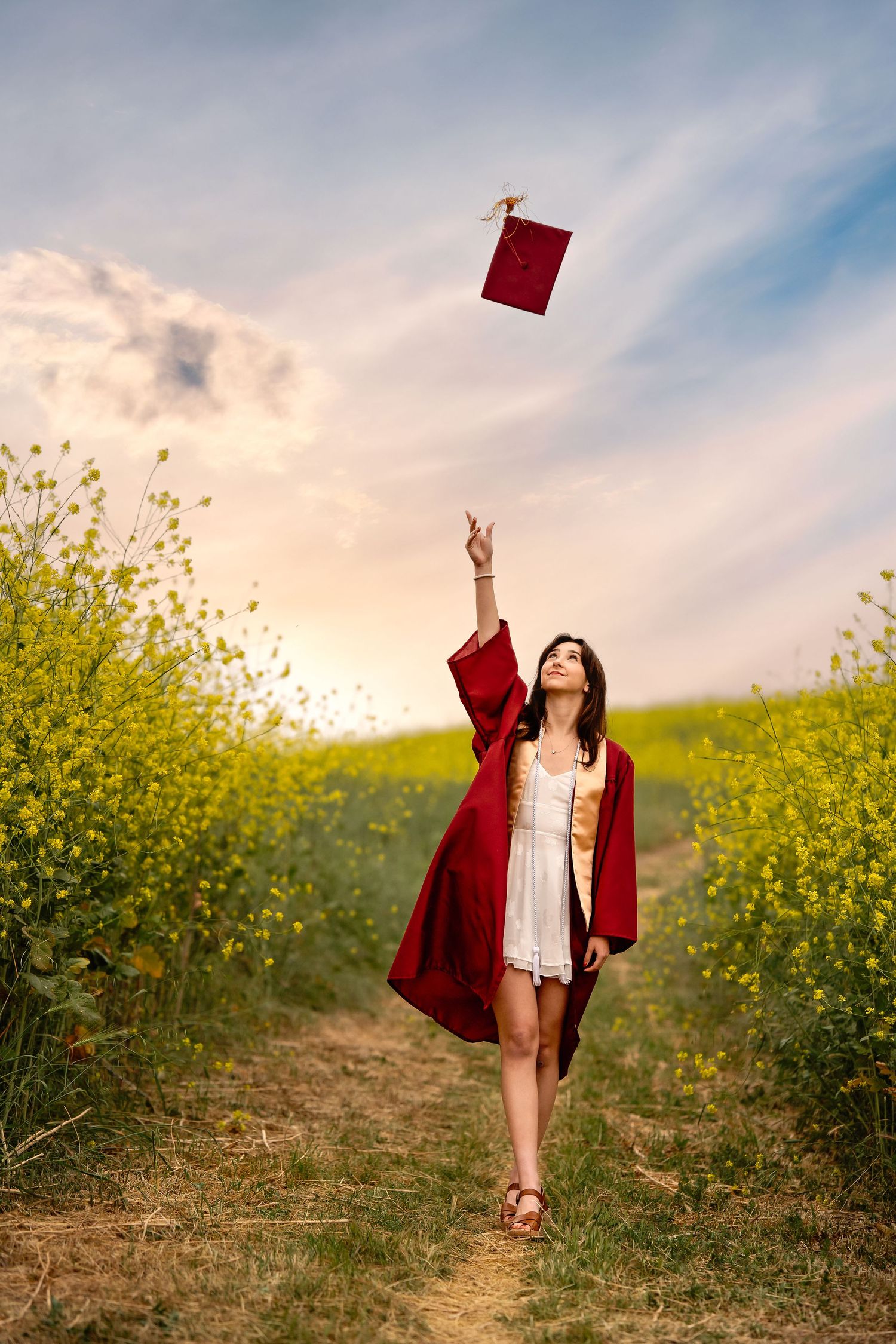 Graduate in red gown celebrating in field of yellow wildflowers at sunset.