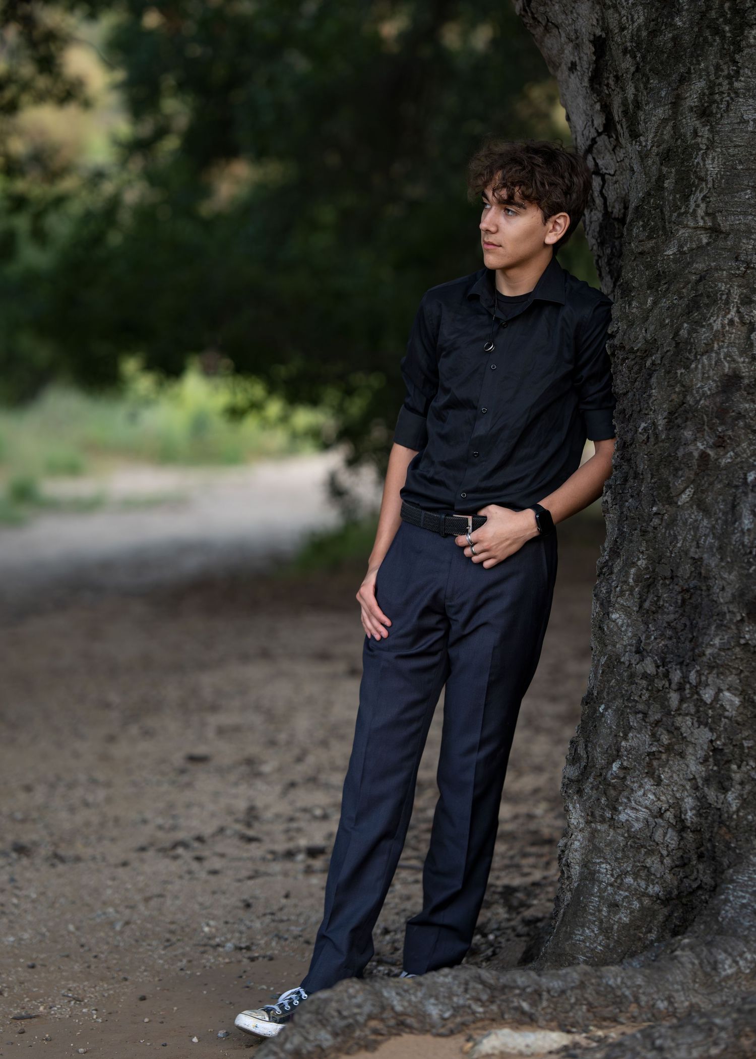 Young man in black shirt and navy pants leaning against a tree on a dirt path in a lush green outdoor setting.