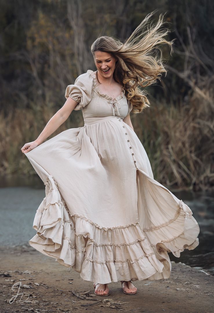 Woman in flowing cream vintage dress spinning on dirt path with wind-blown hair.