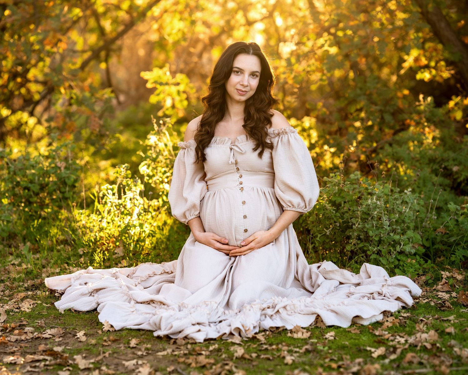 Maternity portrait of woman in white dress sitting among golden autumn foliage.