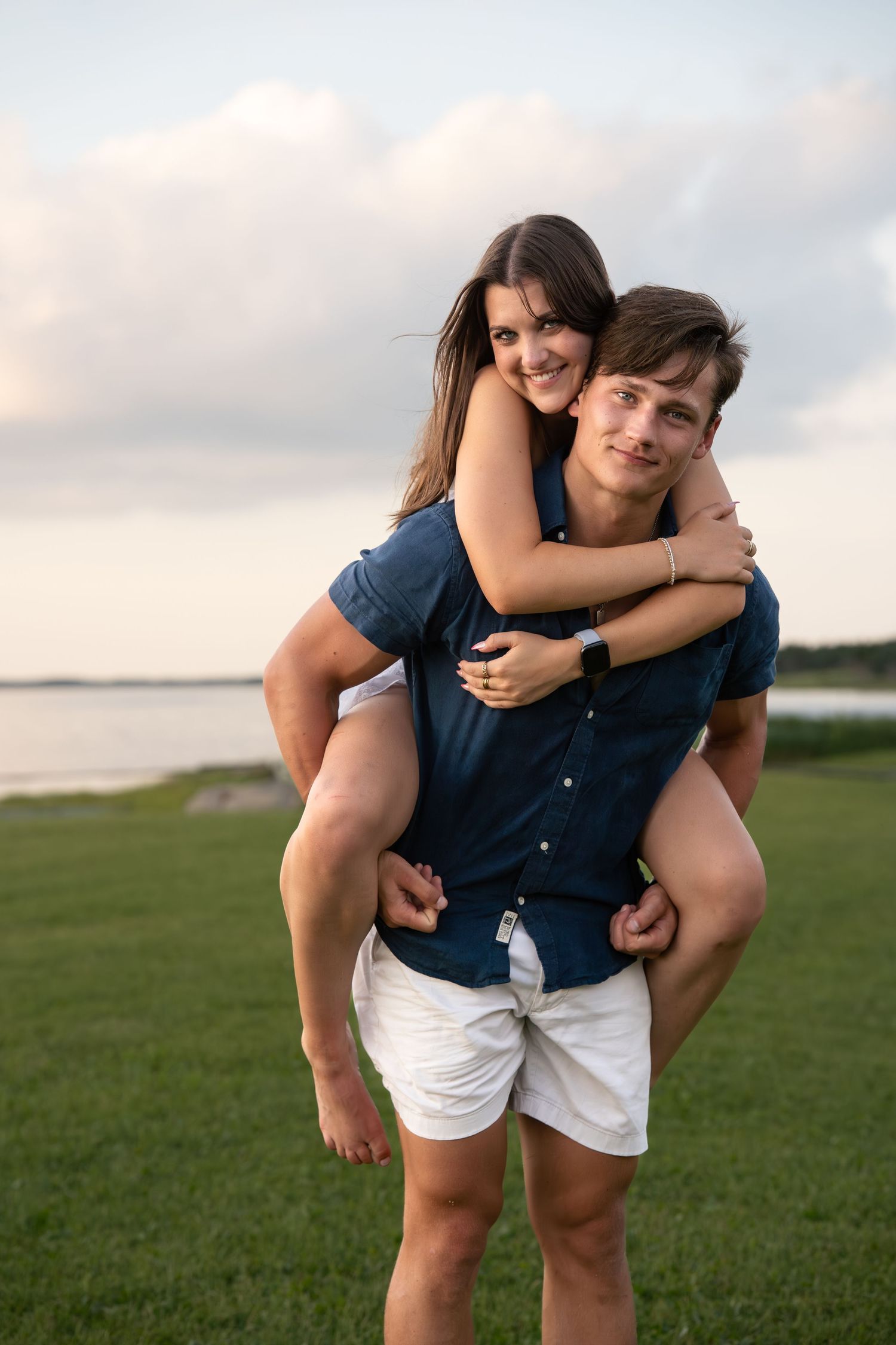 Couple sharing playful piggyback ride moment on green grass by water.