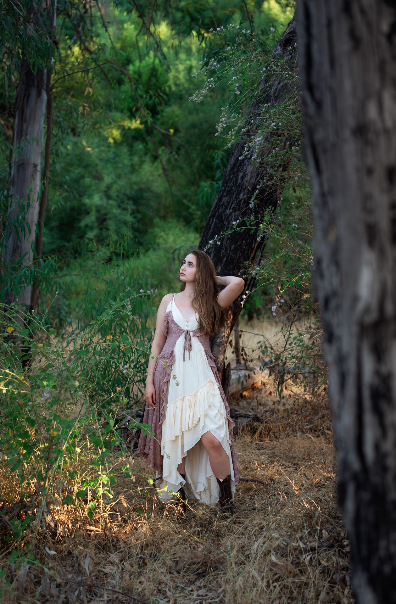 Young woman in a flowing white and mauve bohemian dress standing among trees in a lush green forest.