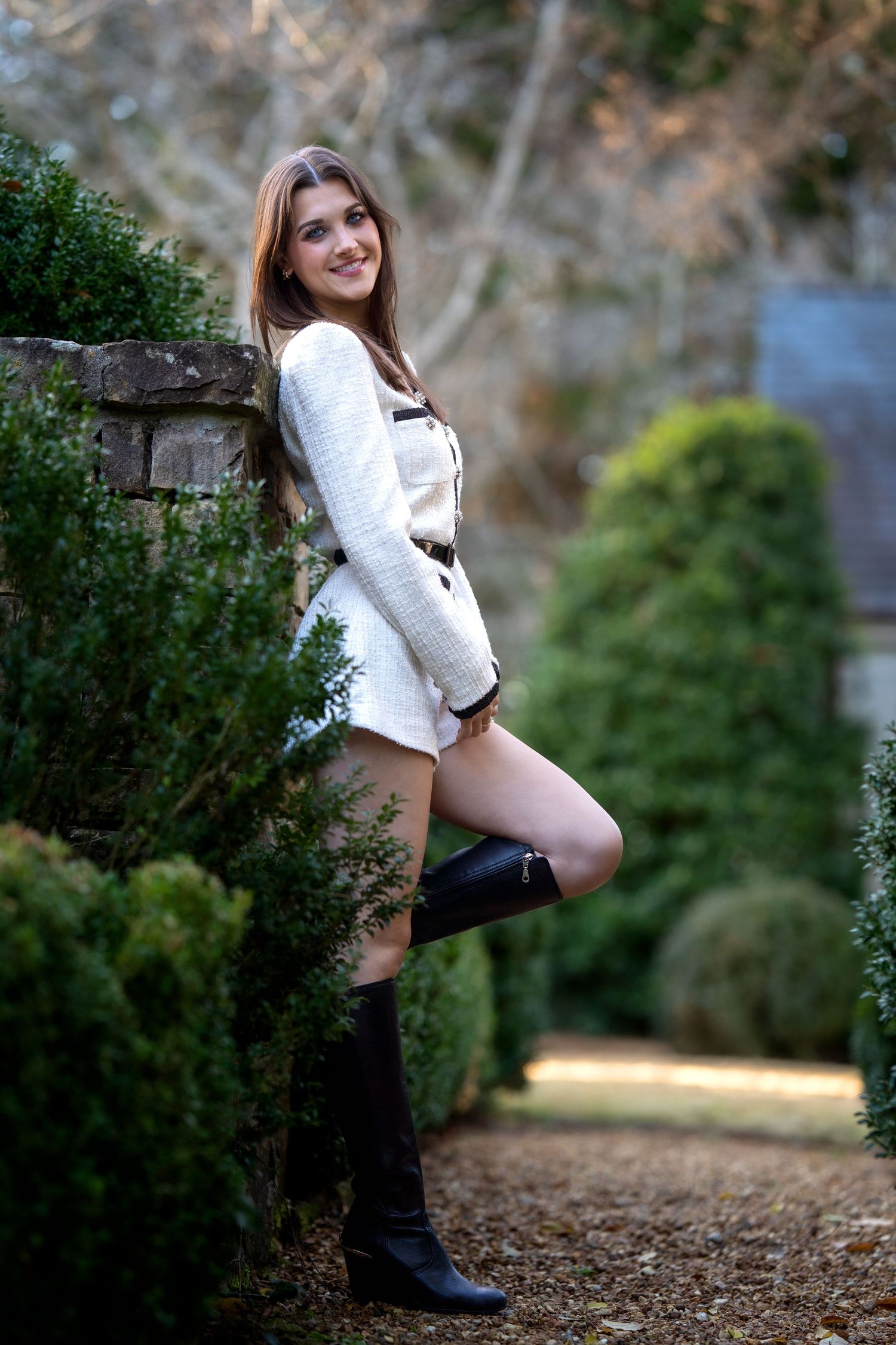 Young woman in white dress and black knee-high boots poses gracefully beside lush green hedges in an outdoor garden setting.