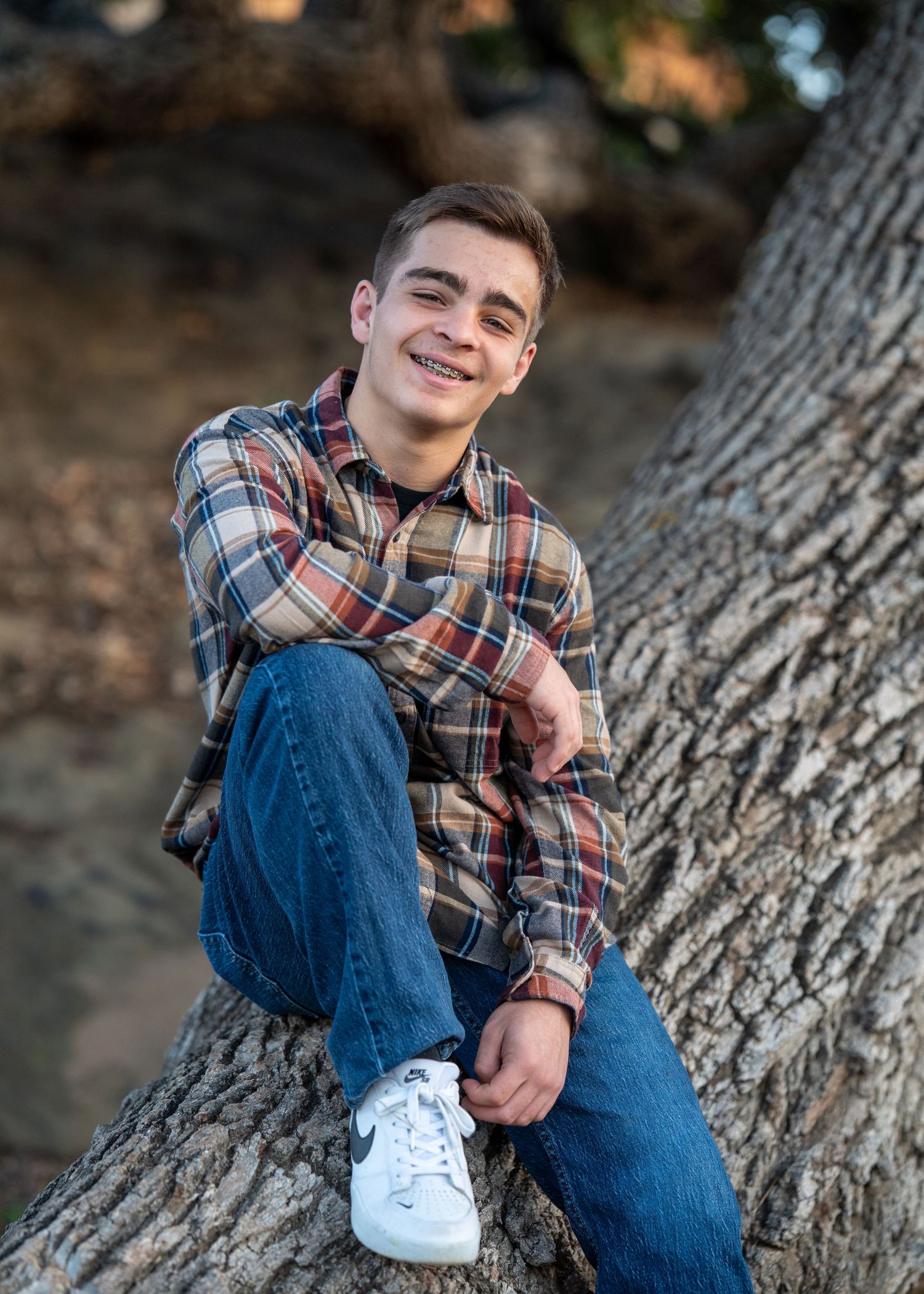 A person in a plaid flannel shirt and jeans sits casually against a large tree trunk outdoors.
