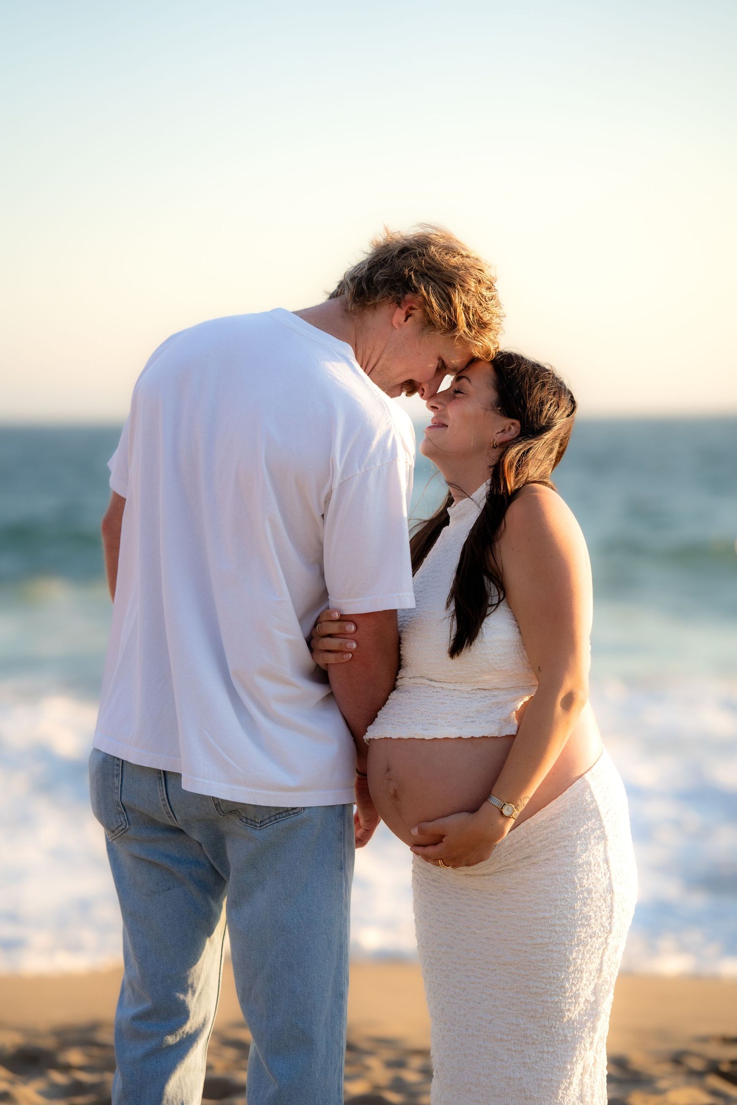 Romantic couple embraces on sandy beach at sunset with ocean waves in background.