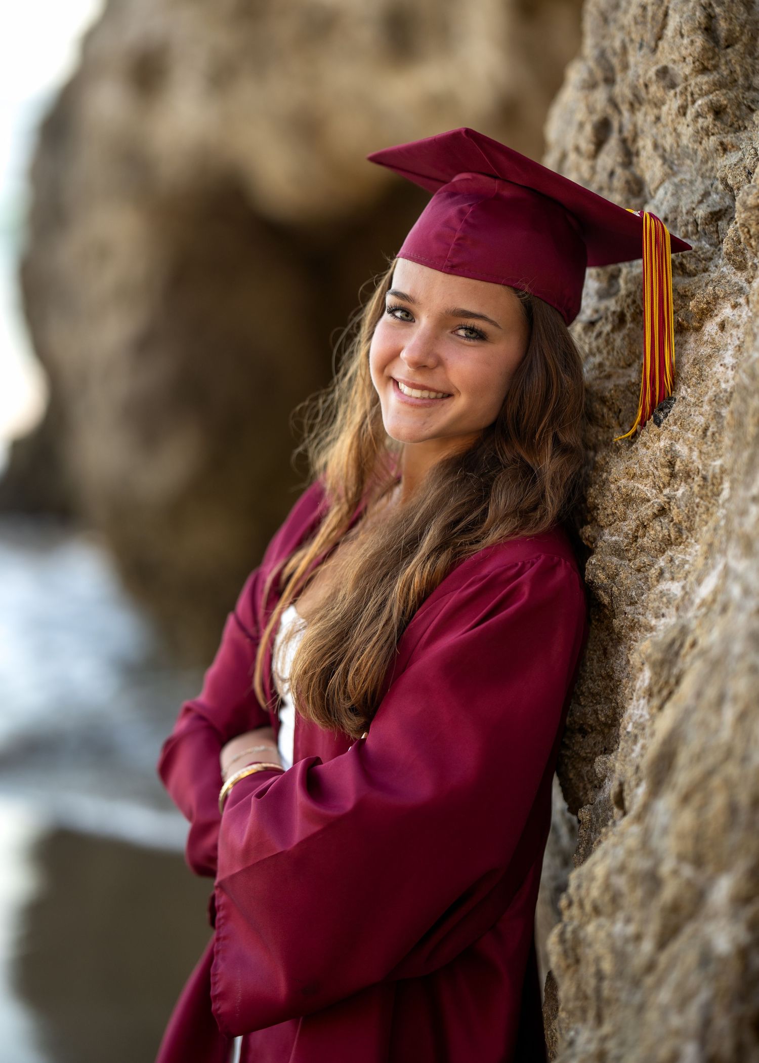 Smiling young woman in maroon graduation cap and gown leaning against rocky coastal cliffs for senior portrait.