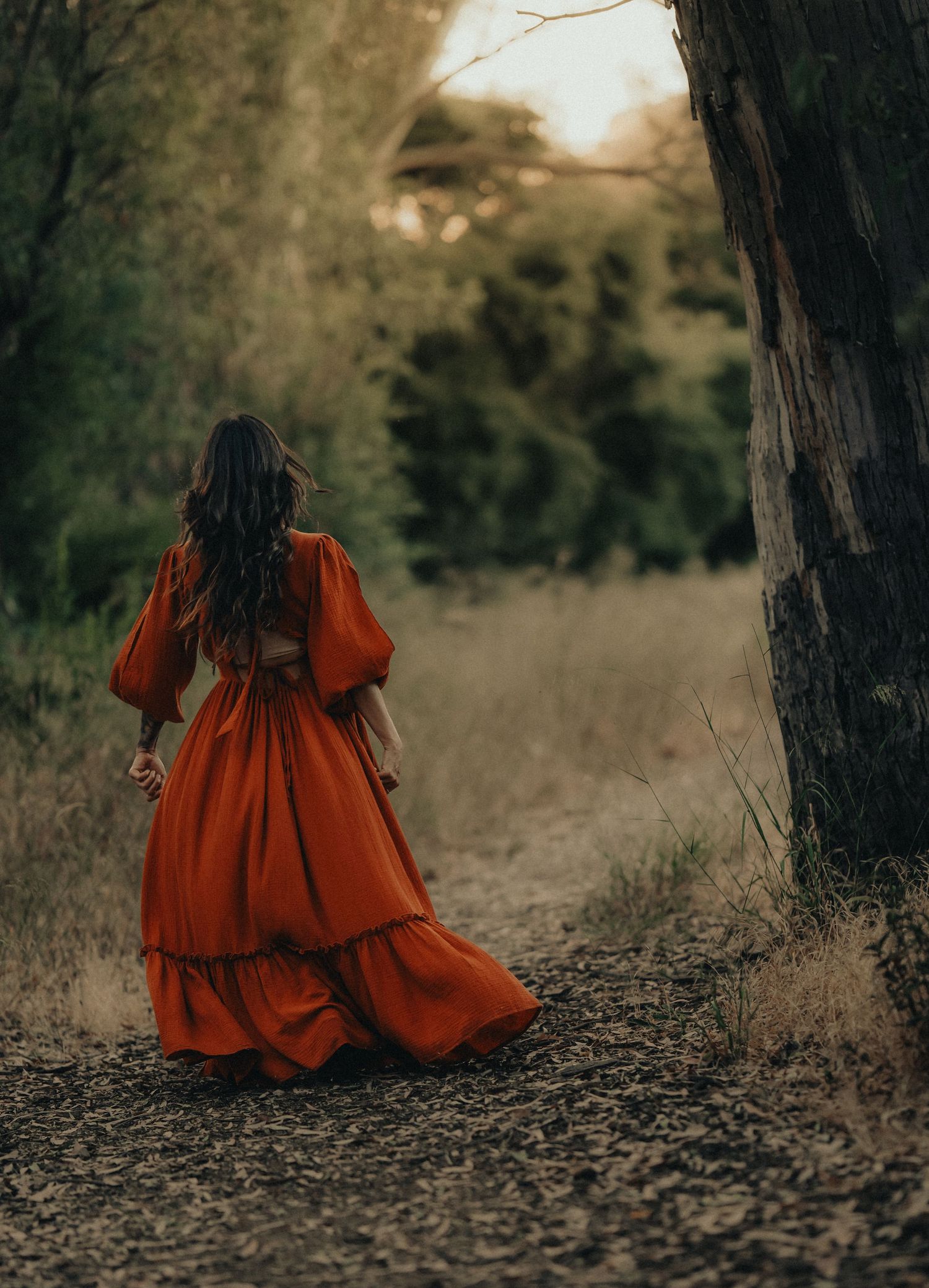 Woman in flowing rust orange dress walks along a wooded path at golden hour.