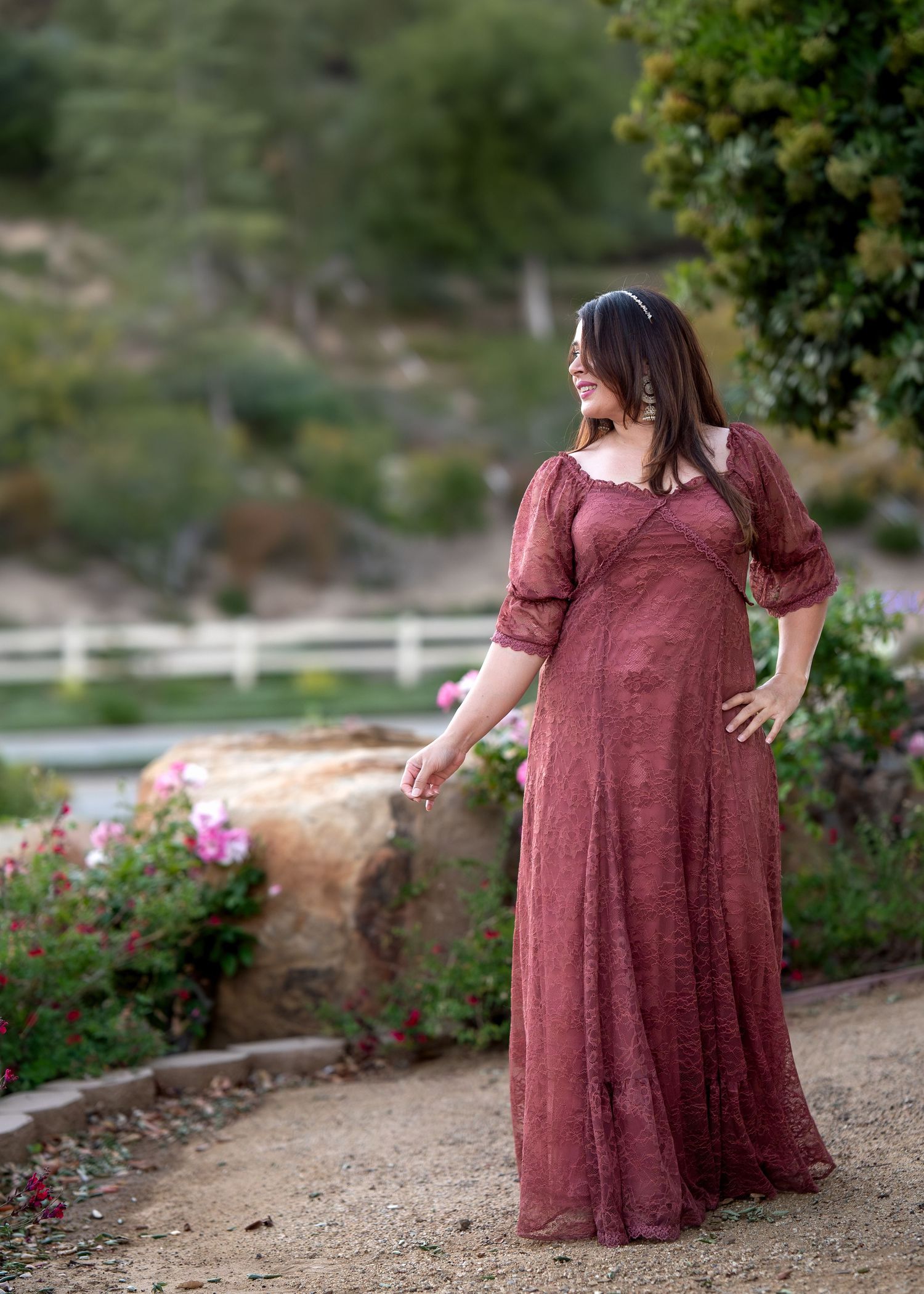 Woman in elegant burgundy lace maxi dress posing outdoors near flowers and white fence.