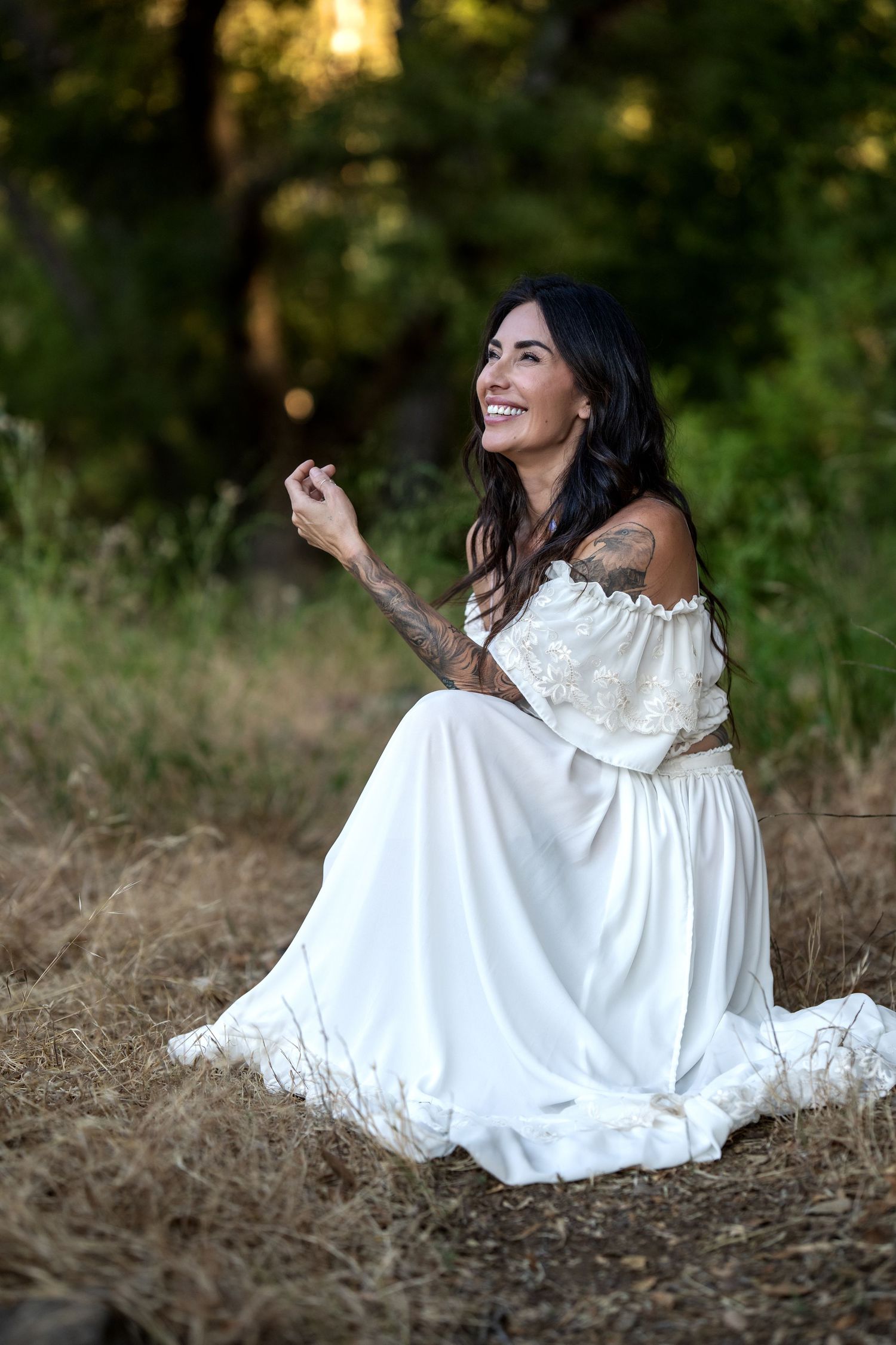 Smiling woman with tattoos in white off-shoulder dress sitting on dry grass in a forest setting.