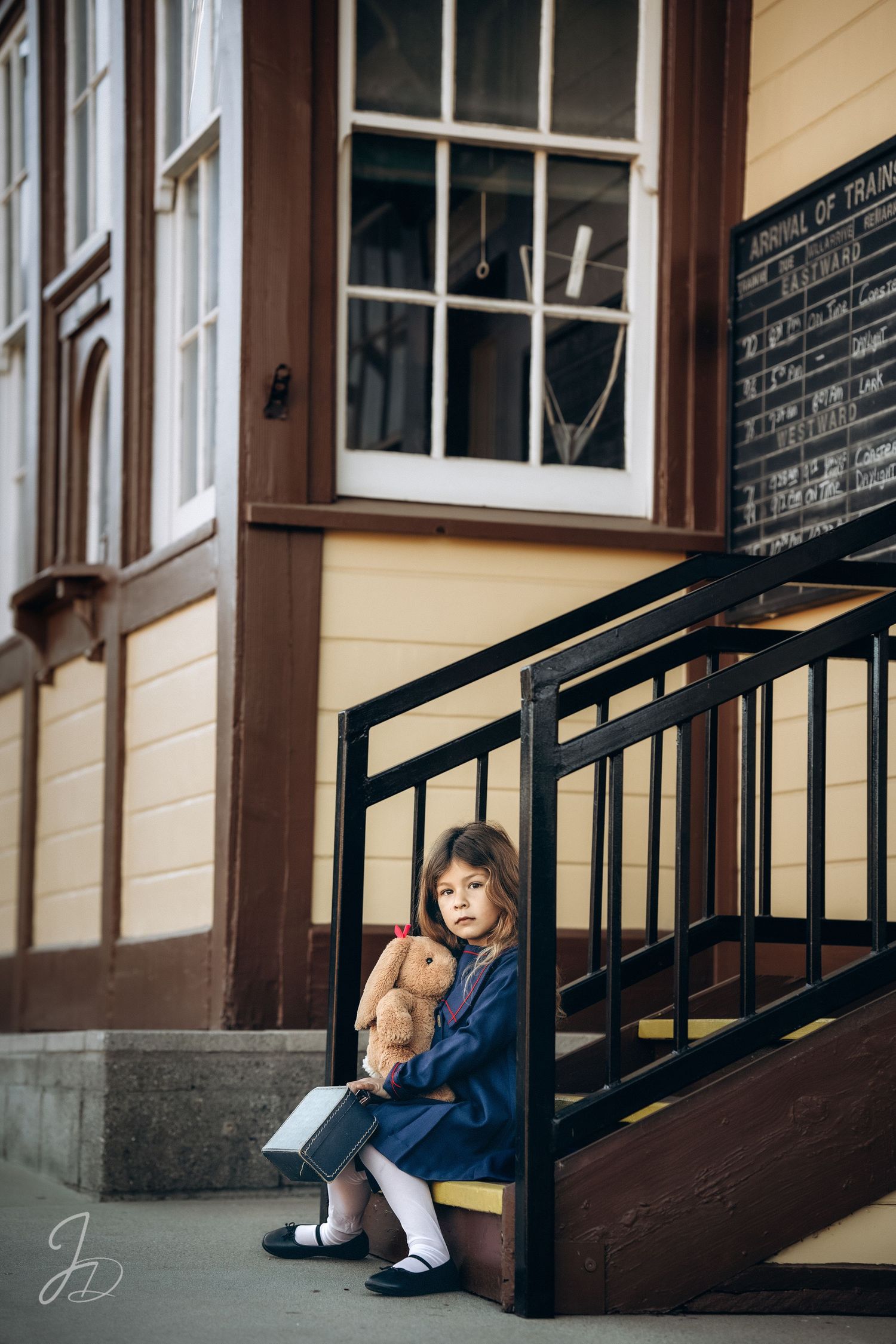 A small figure sits on steps holding a teddy bear outside a vintage building.