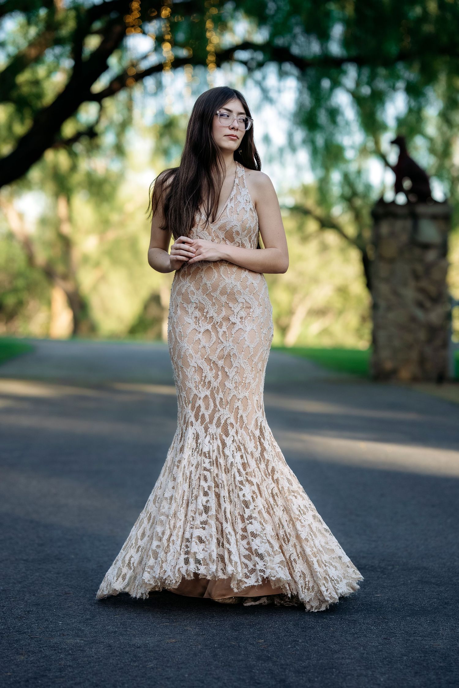 A person in a stunning cream lace mermaid gown poses on a path with soft natural lighting and trees in background.
