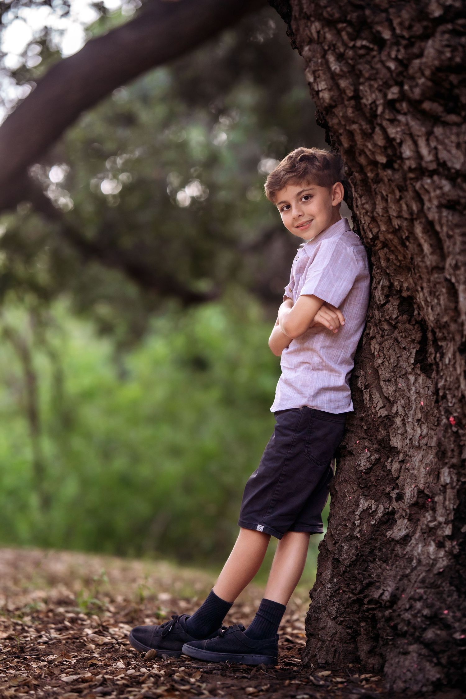 A child leans against a large tree trunk in a park setting wearing a light colored shirt and dark shorts.