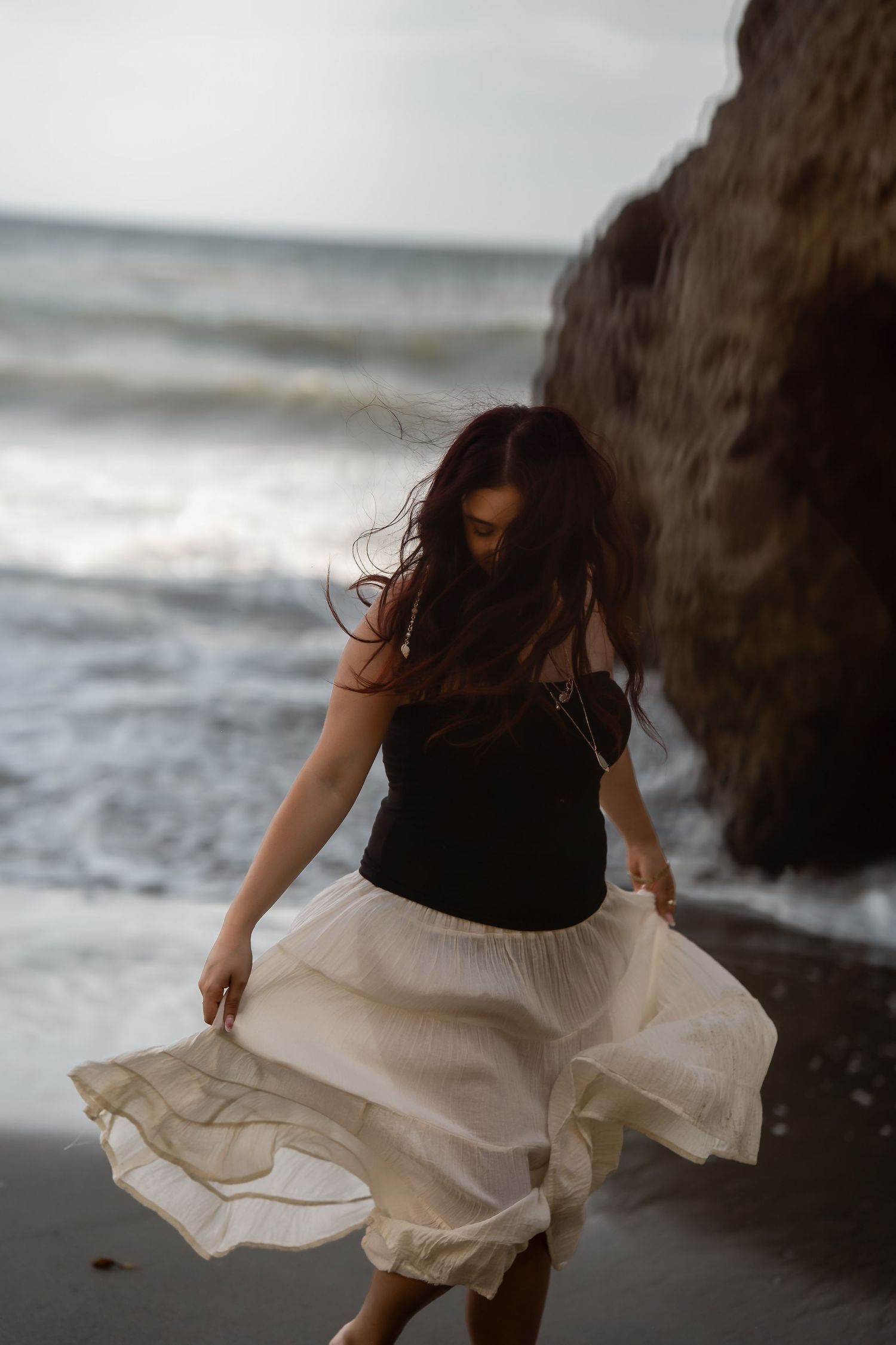 Person twirling in a flowing white dress on a moody beach with rock formations.