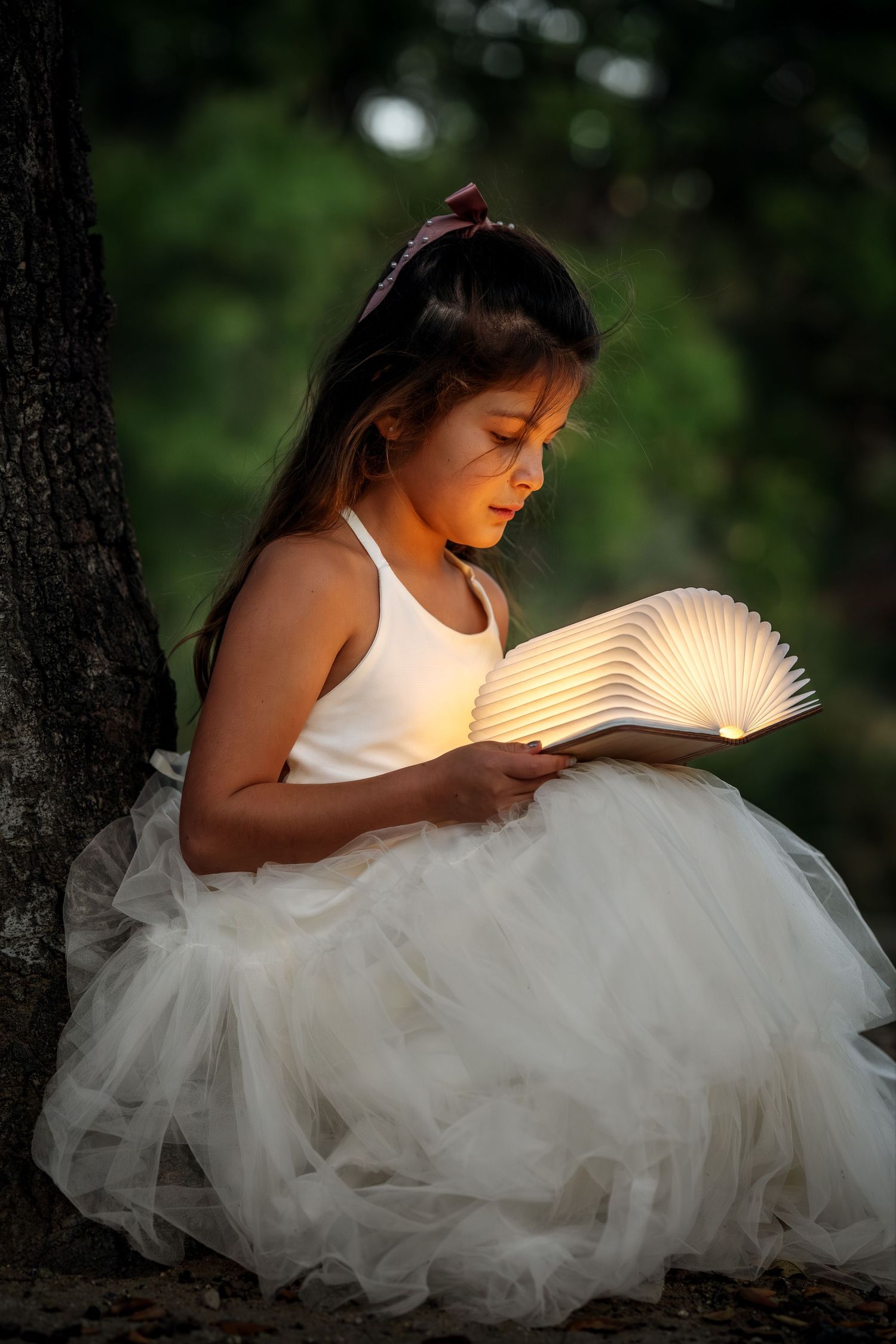 A dreamy portrait of someone in a white tulle dress reading an illuminated book in a forest setting.