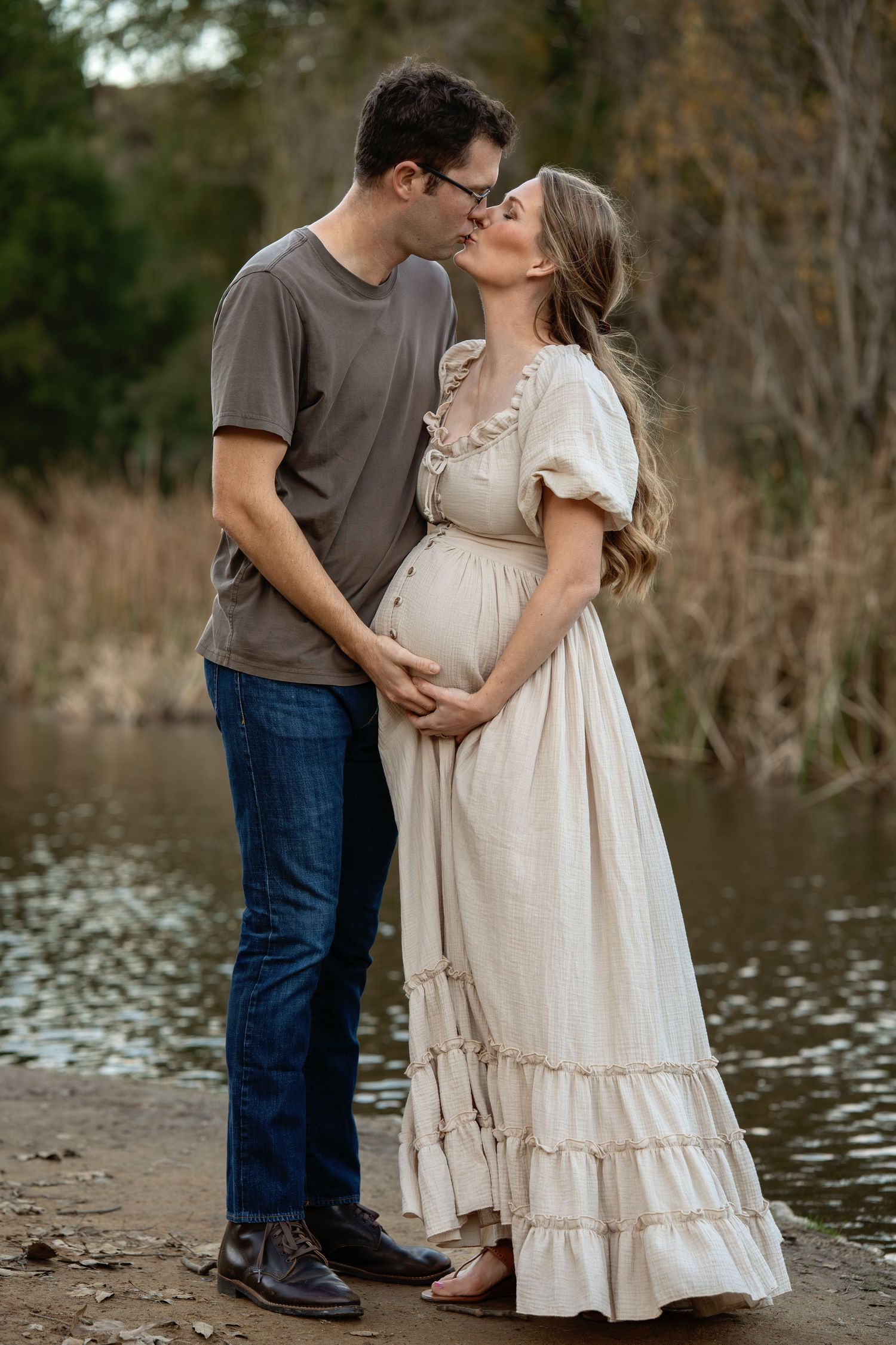Romantic maternity photo session by a lake showing couple embracing in soft natural light.