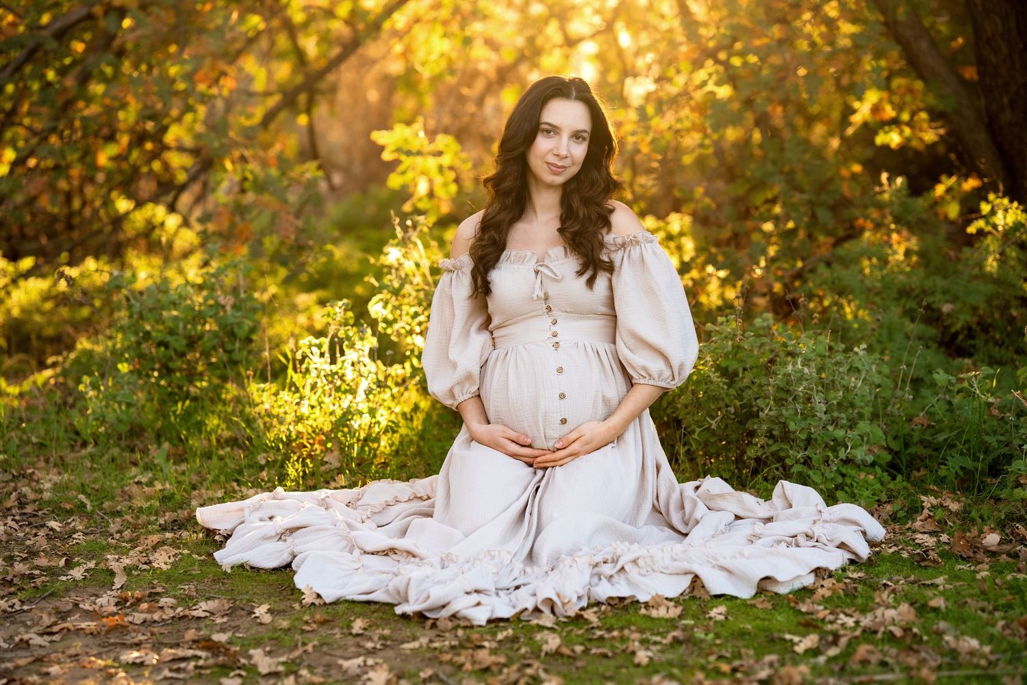 White dress maternity photos in autumn forest with golden sunlight filtering through trees.