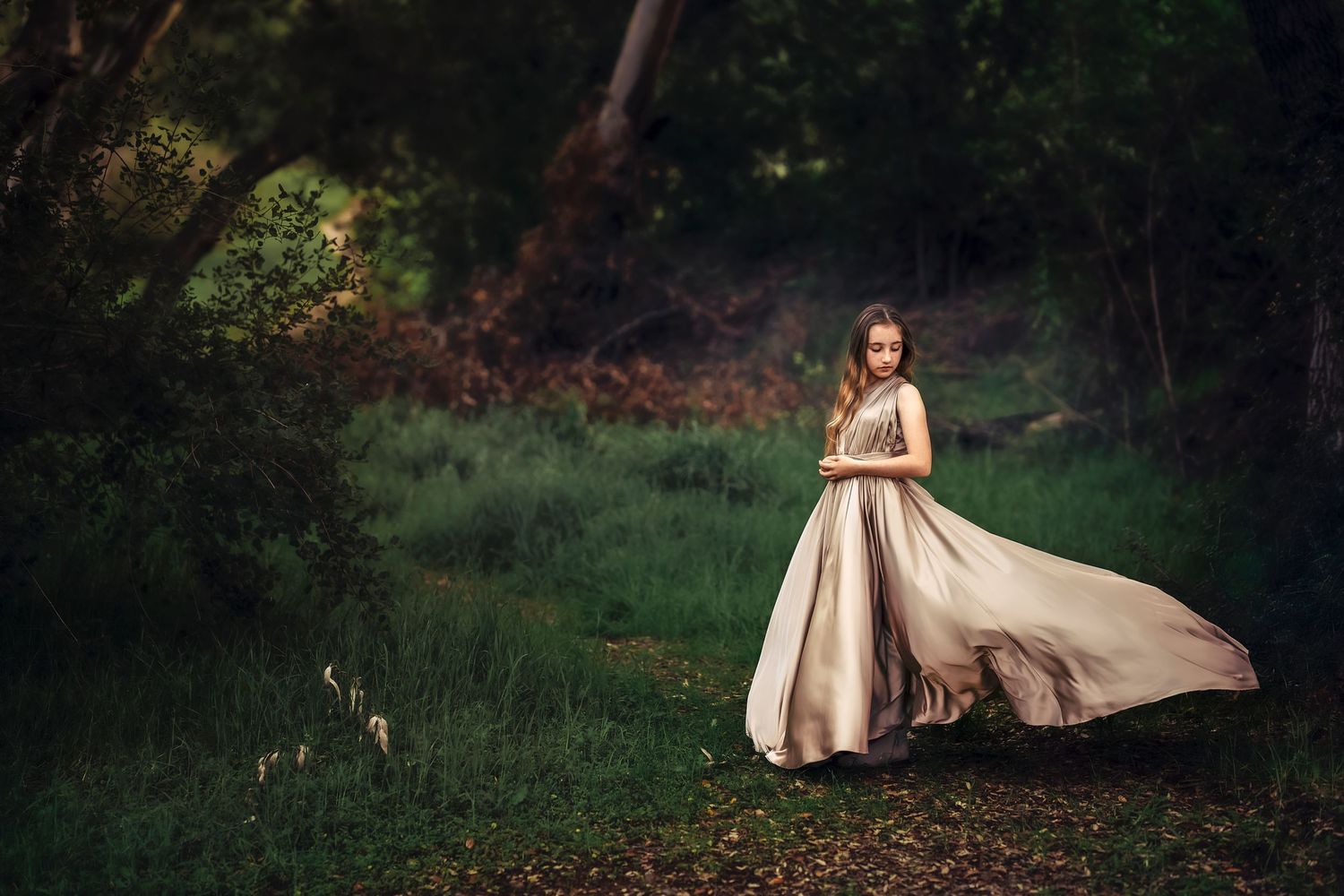 A flowing champagne-colored gown billows elegantly in a mystical forest setting during golden hour.