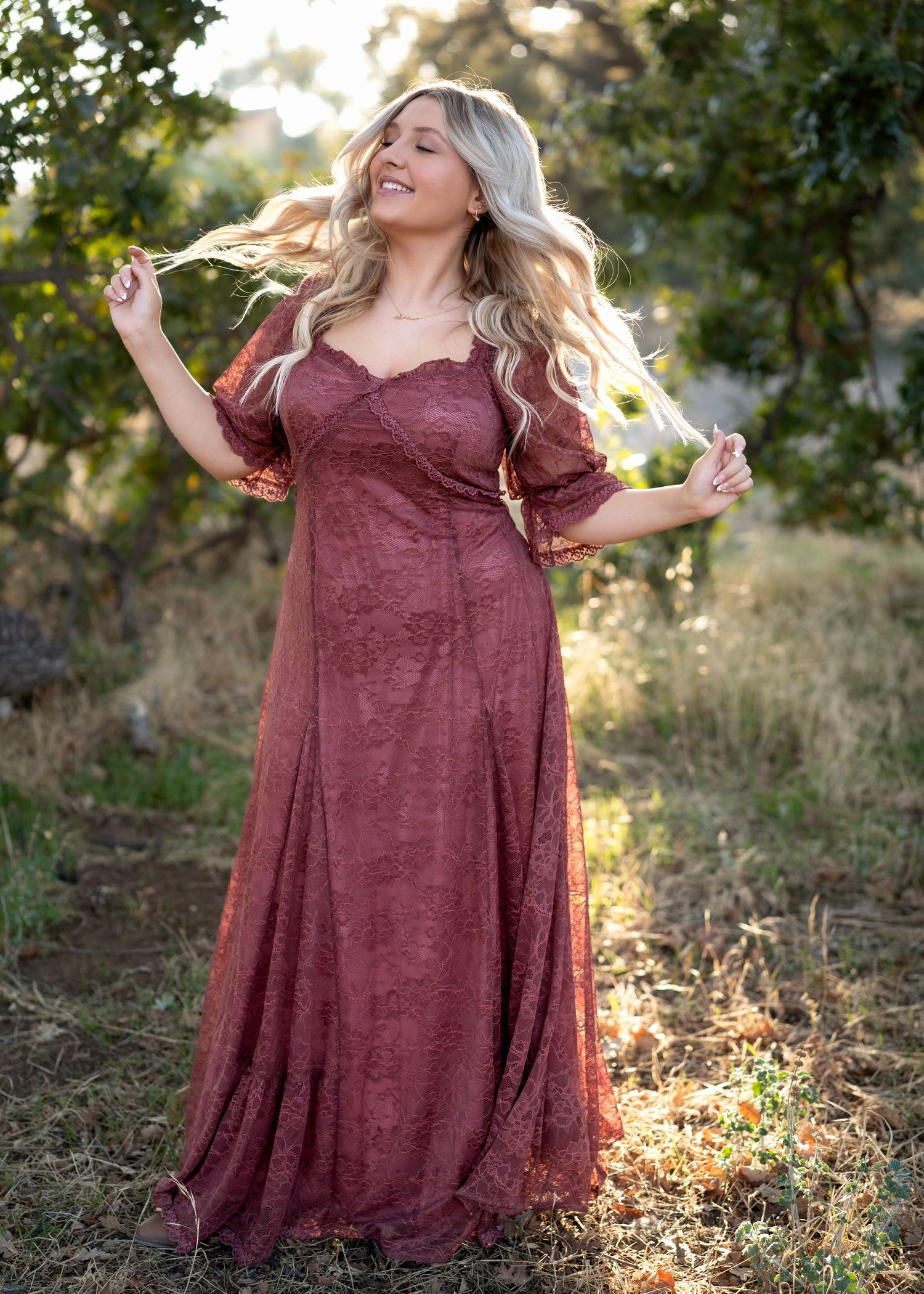 Woman in burgundy dress twirling outdoors with sunlit trees in background.