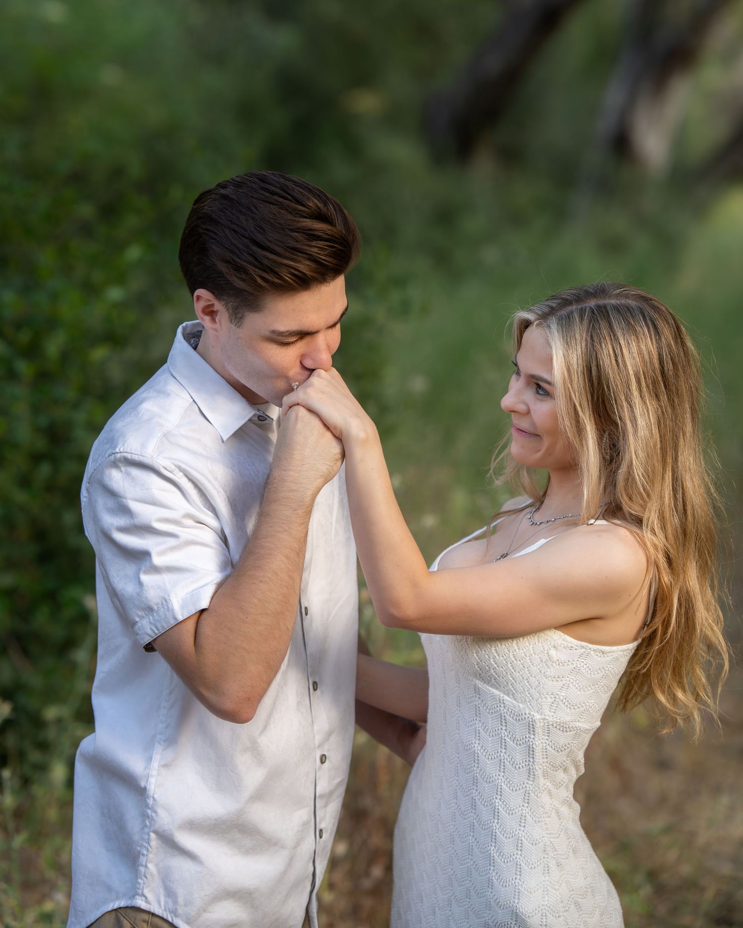 A young man in a white shirt kisses a woman's hand outdoors in a romantic engagement photo session.