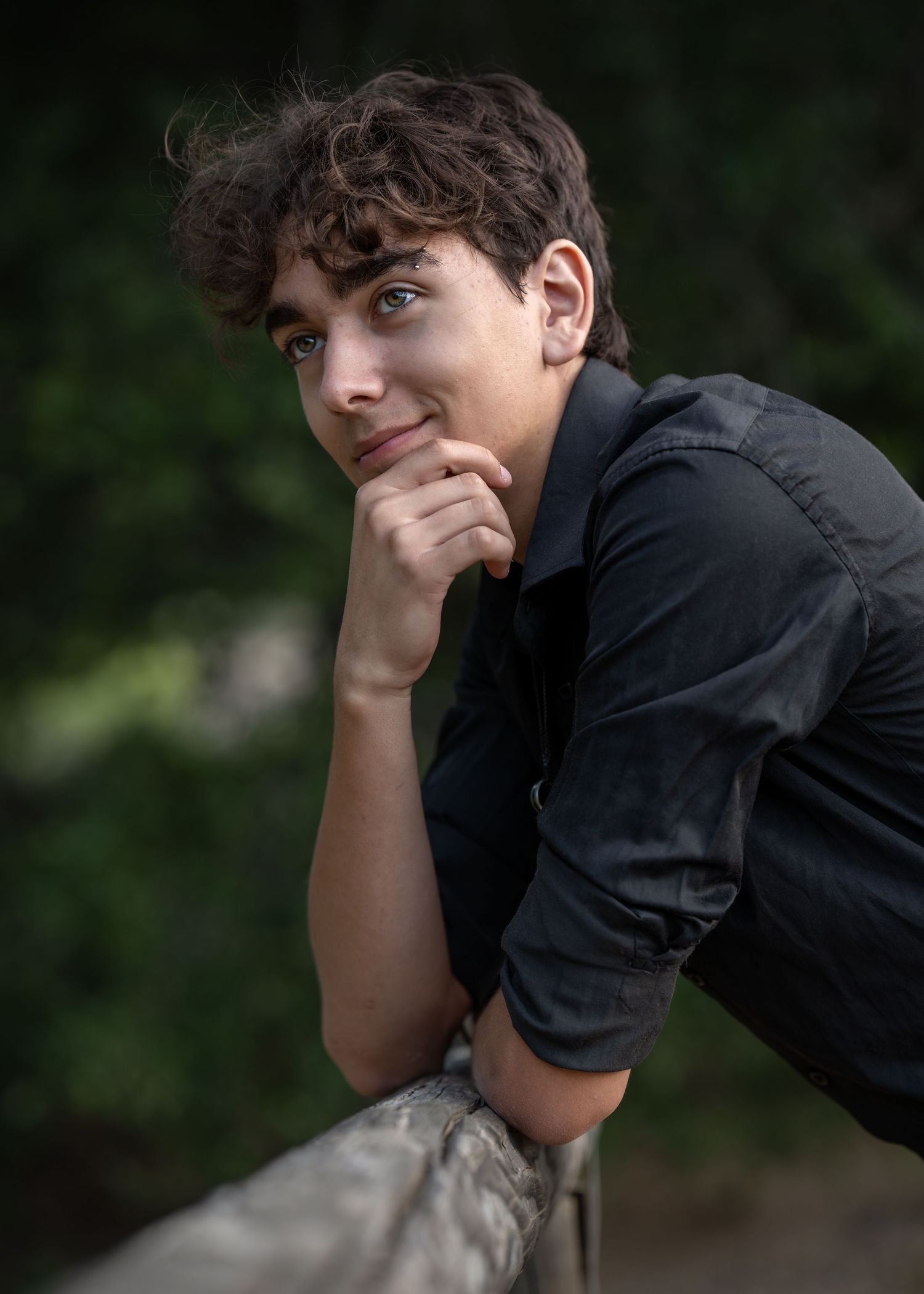 Young man with curly hair in black shirt resting chin on hand, leaning on railing outdoors.