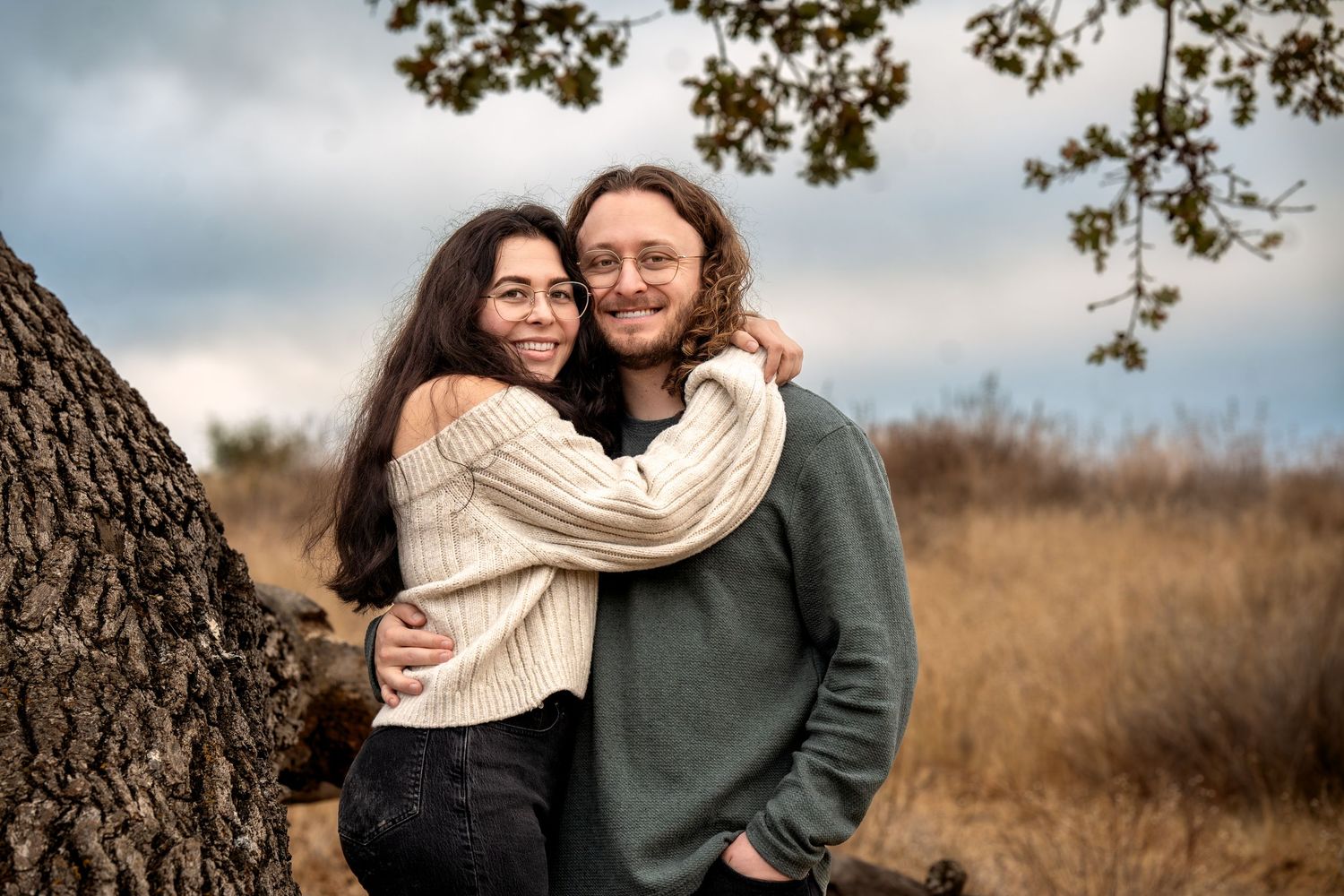 Couple sharing intimate moment against tree in autumn field wearing cozy sweaters.
