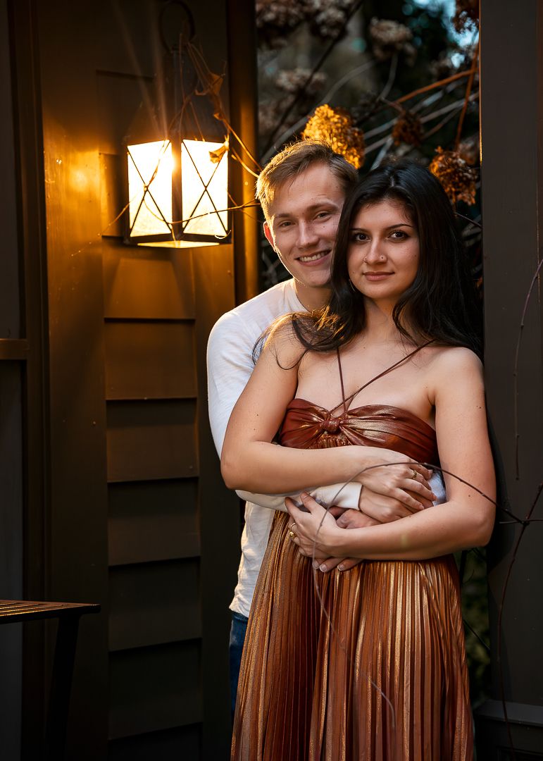 Romantic couple embraces by lantern light outside rustic cabin with woman wearing copper pleated dress.
