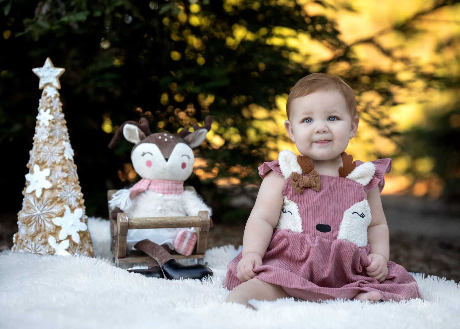 Baby in pink dress sitting among Christmas decorations and artificial snow.