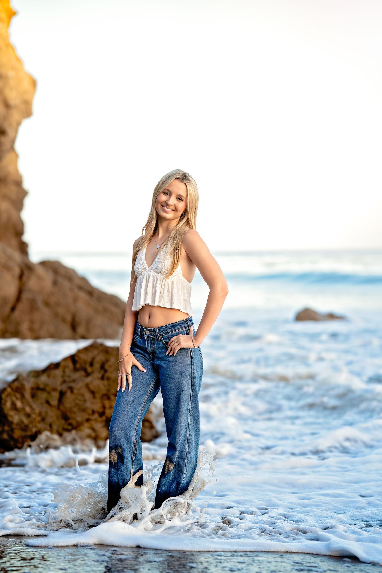 Person in casual attire poses playfully in ocean waves near rocky beach formations.