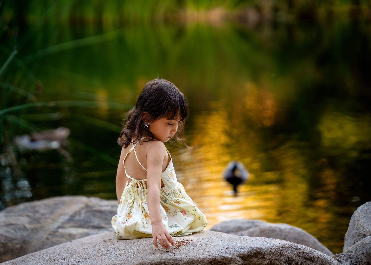 A small figure in white sits contemplatively by a pond with a duck swimming in the golden-lit water.