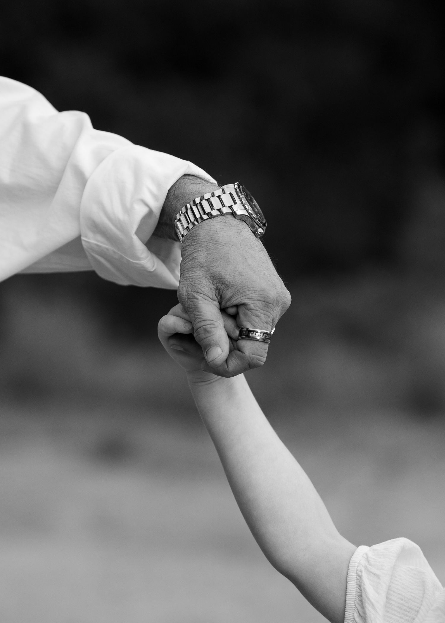 A black and white close-up of an adult hand holding a child's hand, showing a watch and wedding ring.