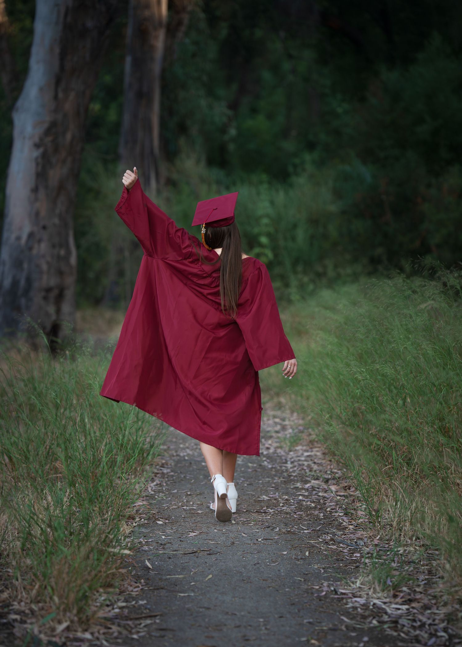 Graduate in red cap and gown walks a forest path, arm raised in celebration.