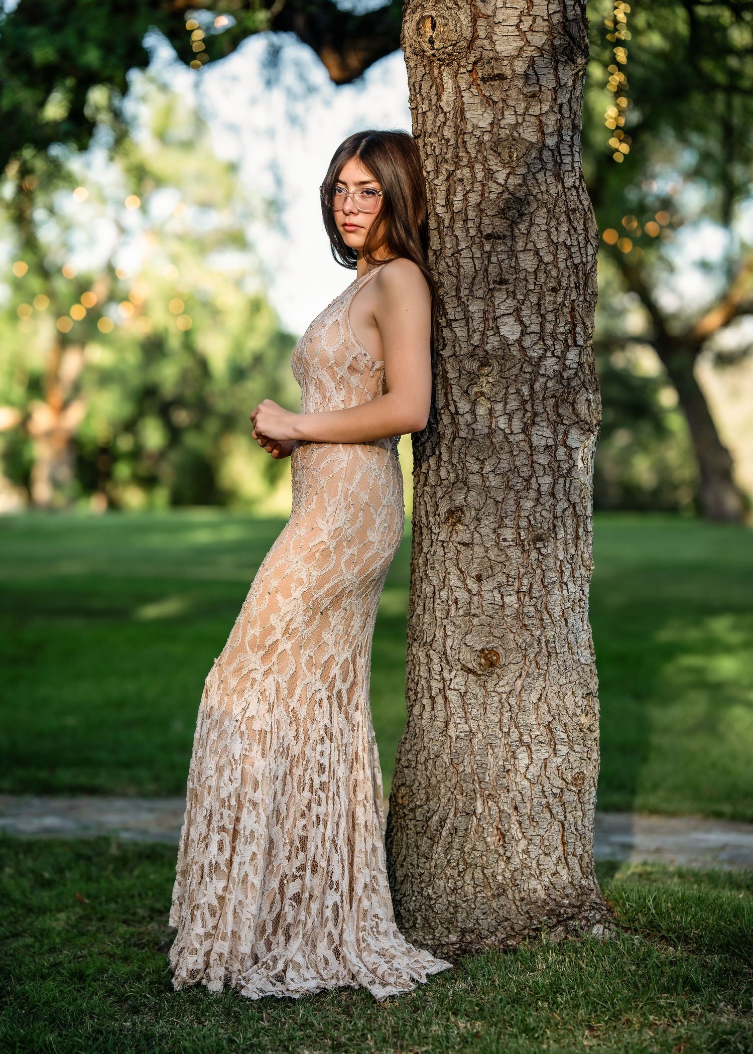 Woman in elegant lace gown leaning against tree trunk in lush green park setting.