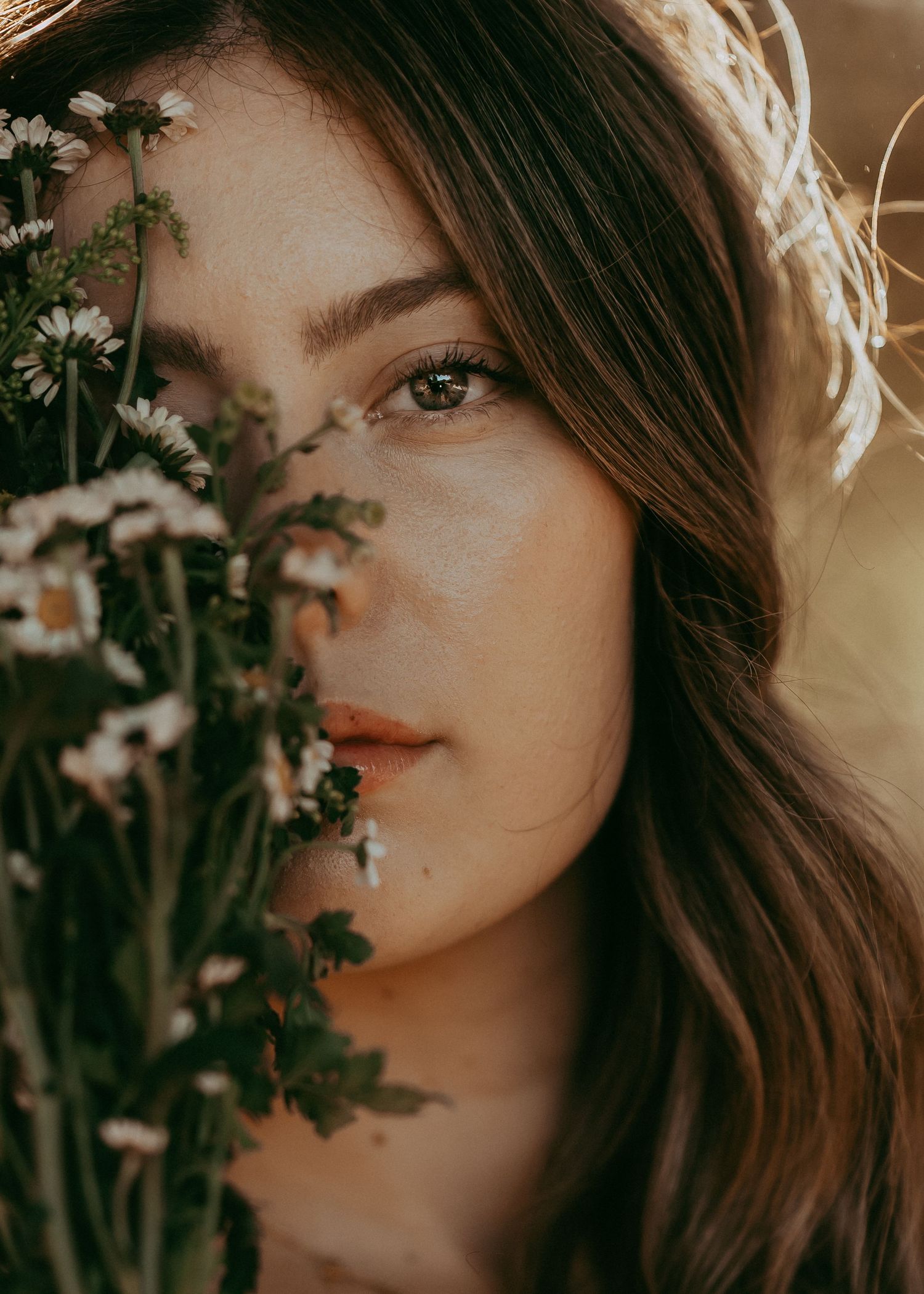 Young woman with brown hair partially hidden behind white daisy flowers in warm golden sunlight.