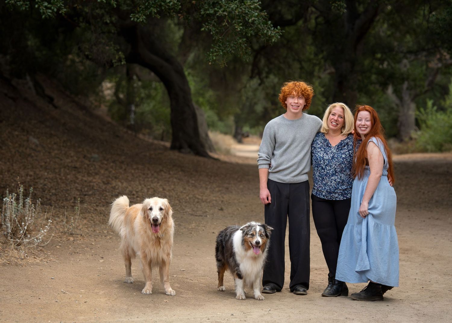 Family of three with red hair poses with two dogs on a shaded dirt trail in a scenic outdoor setting.